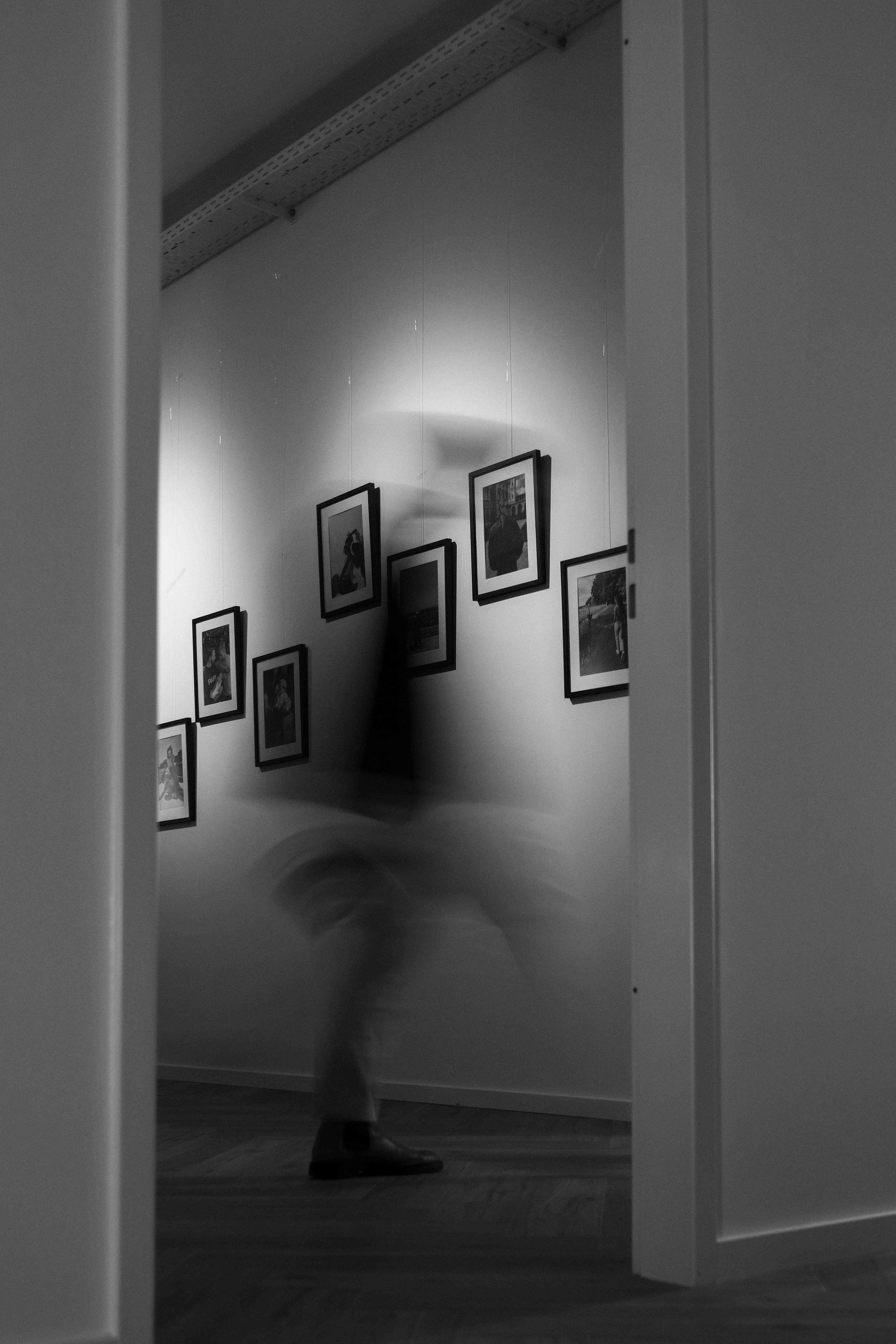 Schwarz-weiß-Fotografie einer Person, die einen Flur in einem Kunstmuseum oder einer Galerie durchquert, mit mehreren an der Wand aufgehängten Bilder. Die Person ist unscharf, was Bewegung andeutet, und der Raum ist minimalistisch gestaltet.