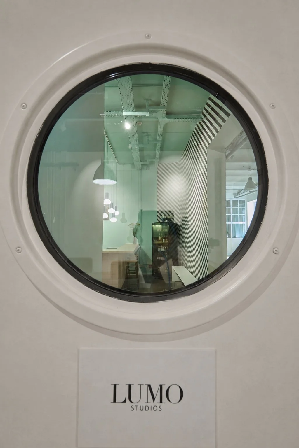 Blick durch das runde Fenster in das farbenfrohe Interior von Lumo Studios München mit Küche, Bar und grafischer Wandgestaltung.
