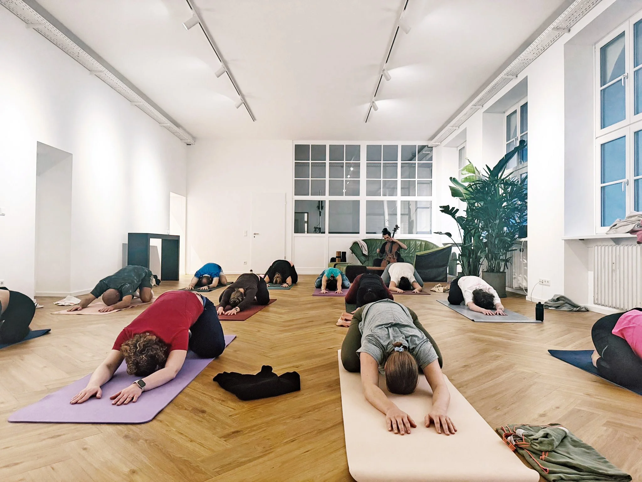 Menschen beim Yoga in einem hellen, modernen Raum, einige in Child's Pose, ein Musiker spielt auf einem Cello im Hintergrund.
