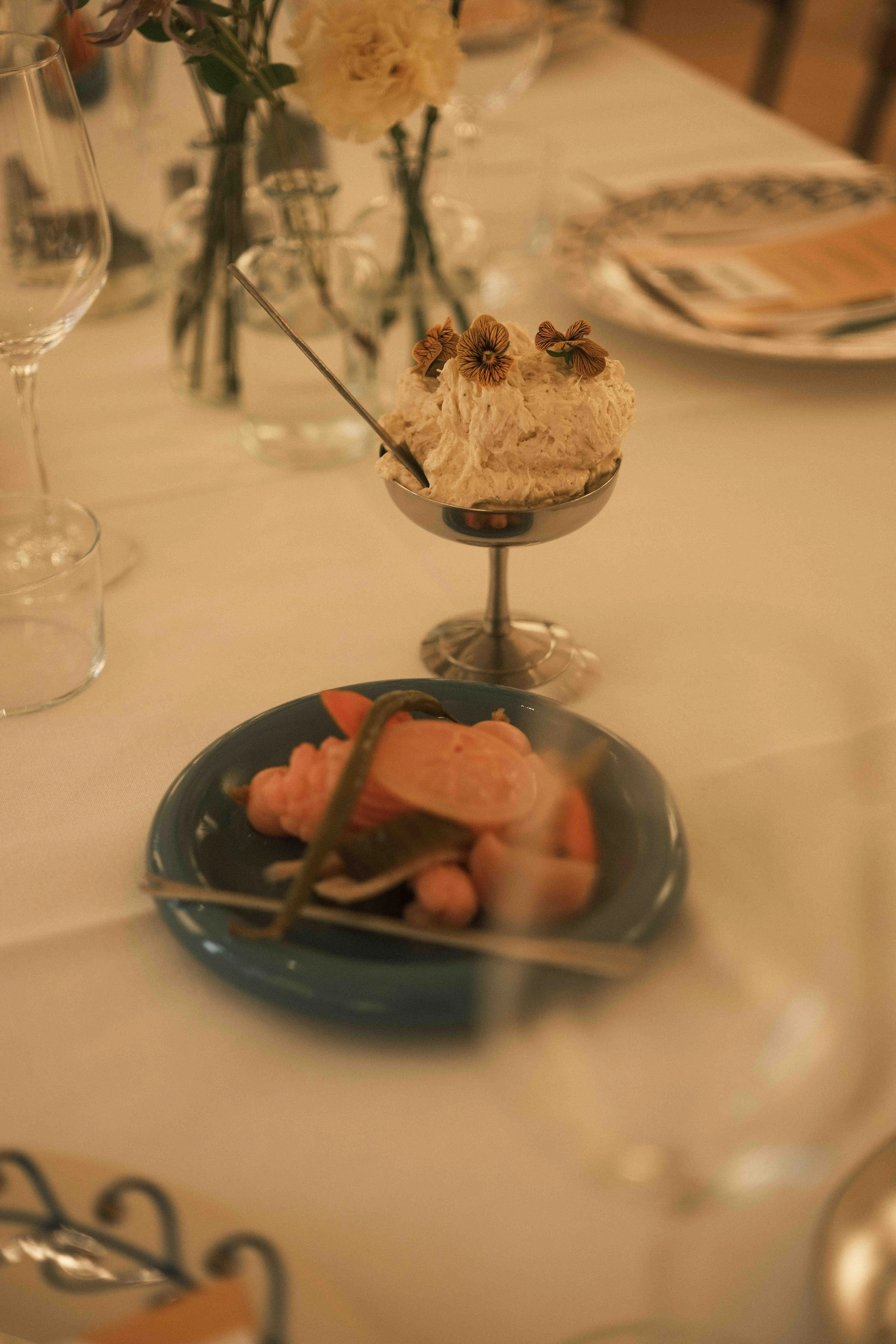 Ein Dessert in einer eleganten Schale mit Blumen, daneben ein Teller mit roter Fisch, Gemüse und Zitronenscheibe auf einem gedeckten Tisch.