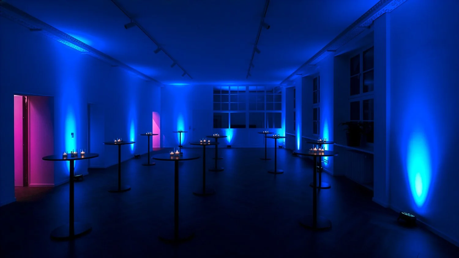 Ein Dunkelraum mit blauer und pinker Beleuchtung, mehreren Stehtischen mit Kerzen, und großen Fenstern an der Wand.