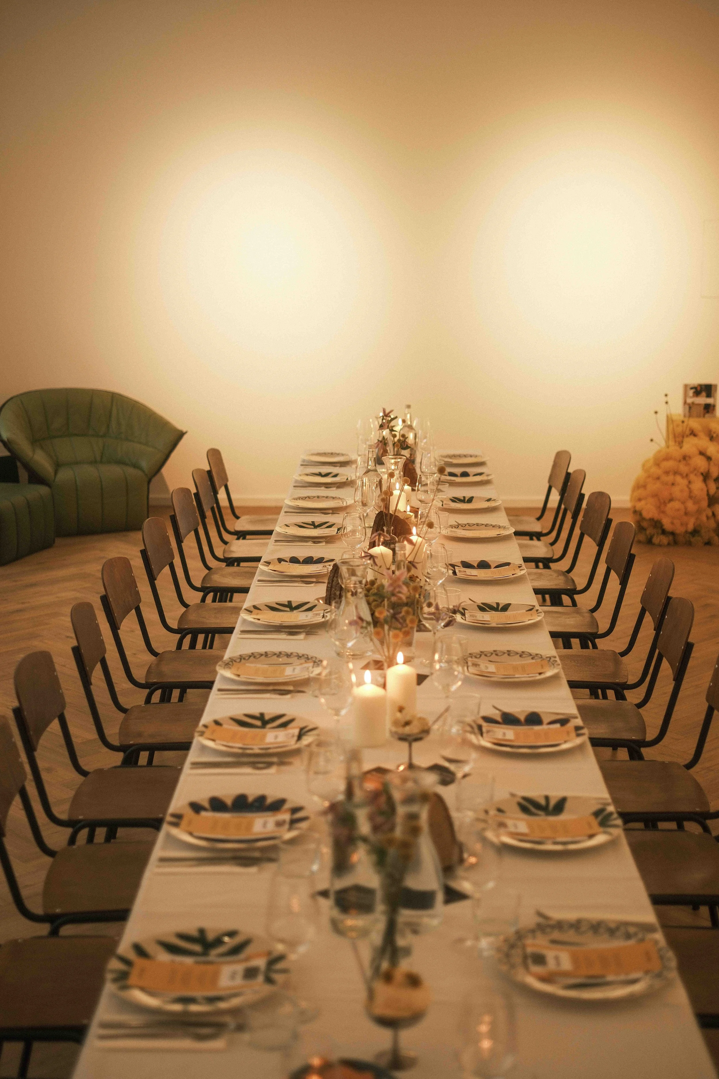 Lange Tafel mit weißen Tischdecke, Wein- und Wassergläsern, Tellern mit grünen und blauen Mustern, Kerzen und Blumen, in einem festlich dekorierten Raum.
