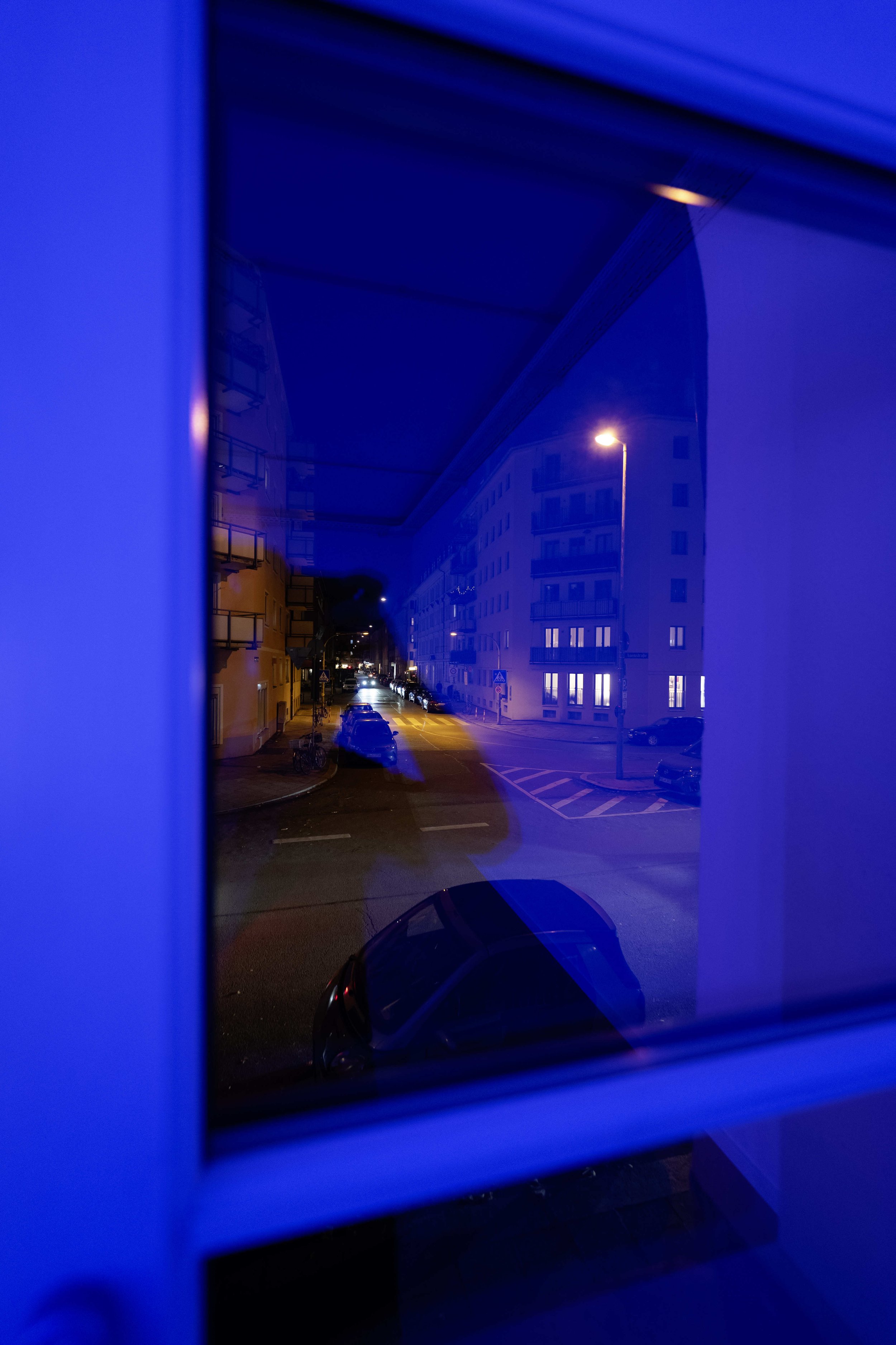 Blick auf eine nächtliche Straße, gesehen durch ein Fenster mit blauer Beleuchtung, mit parkenden Autos und beleuchteten Gebäuden.