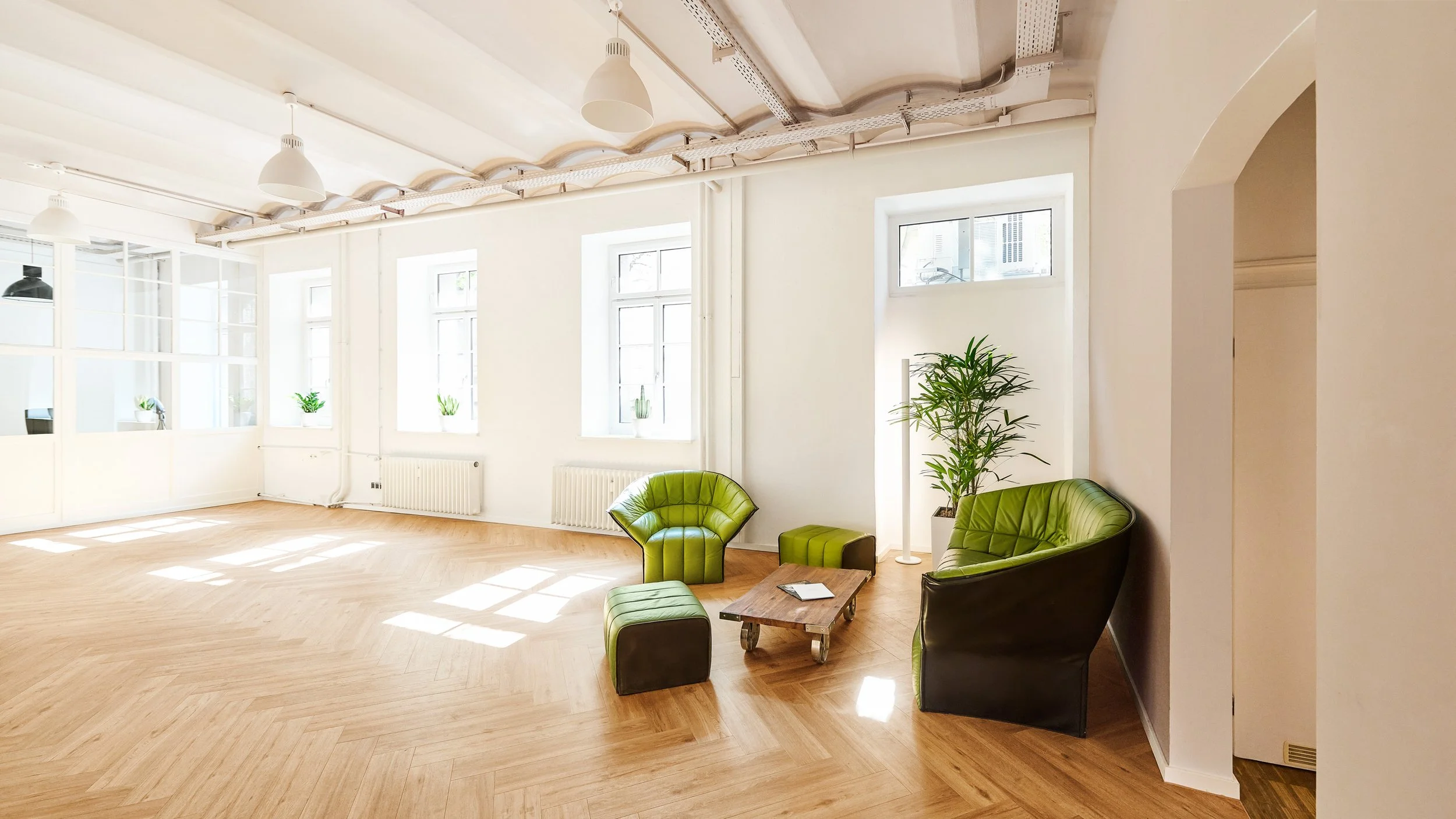 Ein helles, modern eingerichtetes Wohnzimmer mit großen Fenstern, grünen Sitzmöbeln, einer kleinen Holztisch und Pflanzen.