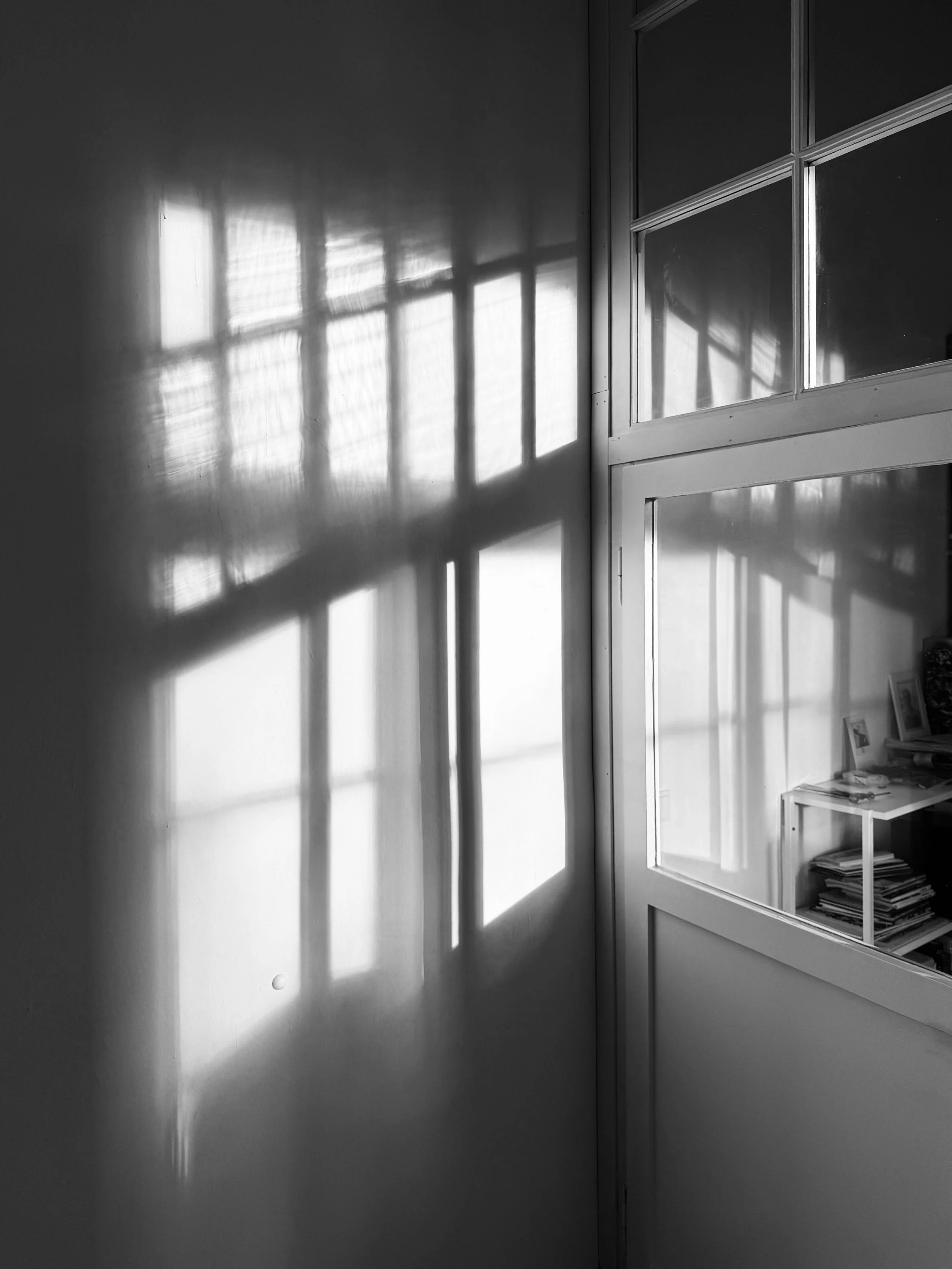 Schwarze und weiße Fotografie eines Fensters mit Gitter, das Sonnenlicht auf die Wand und ein Regal mit Büchern und Fotos projiziert.
