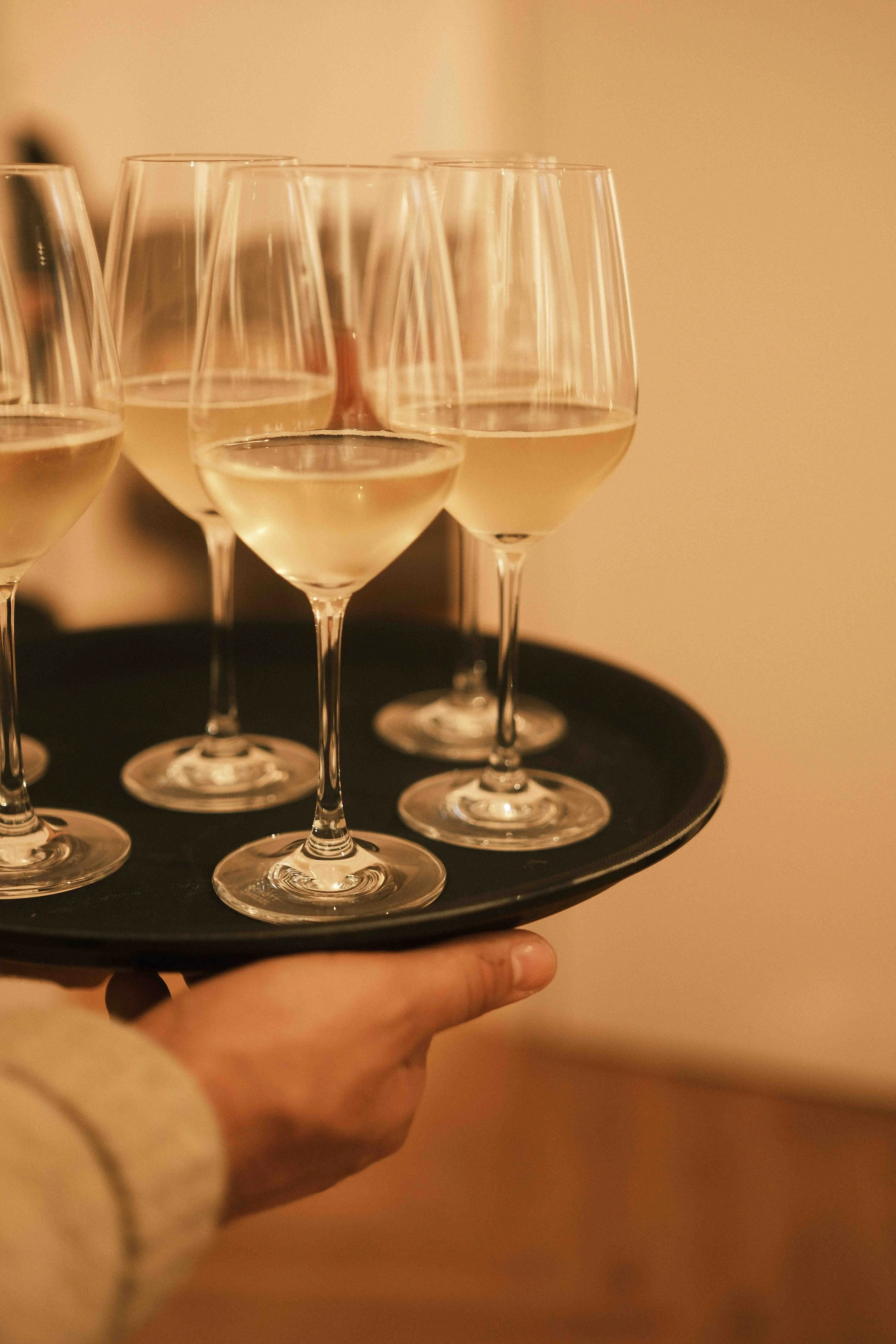 Hand hält einen schwarzen Teller mit mehreren weißen Wein gläsern, gefüllt mit Weißwein, in einem warm beleuchteten Raum.