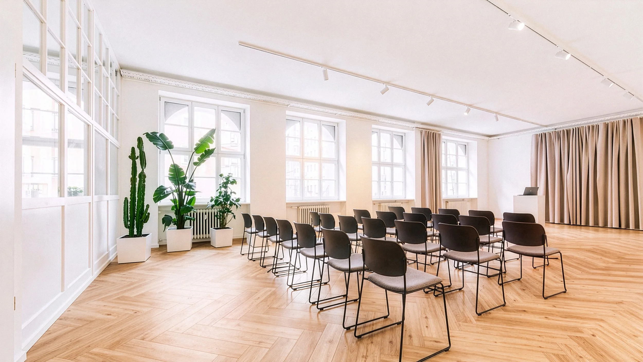 Ein heller, moderner Seminarraum mit großen Fenstern, mehreren schwarzen Stühlen, Pflanzen und einem Vortragspult.