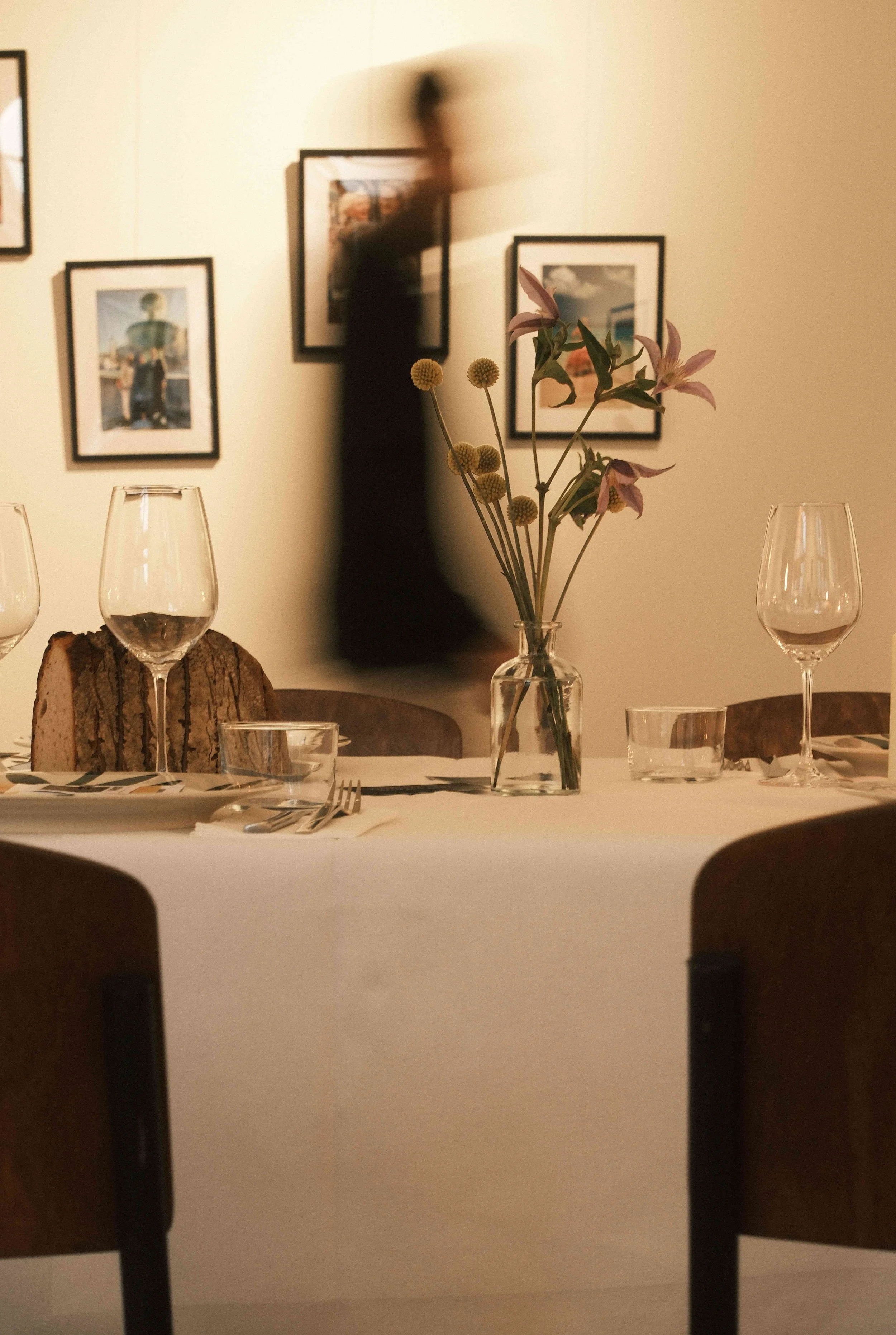 Ein festlich gedeckter Tisch mit Weingläsern, einer Vase mit Blumen, einem Brotlaib und Geschirr, im Hintergrund eine Wand mit Framed-Artworks und eine schwebende, verschwommene Figur.
