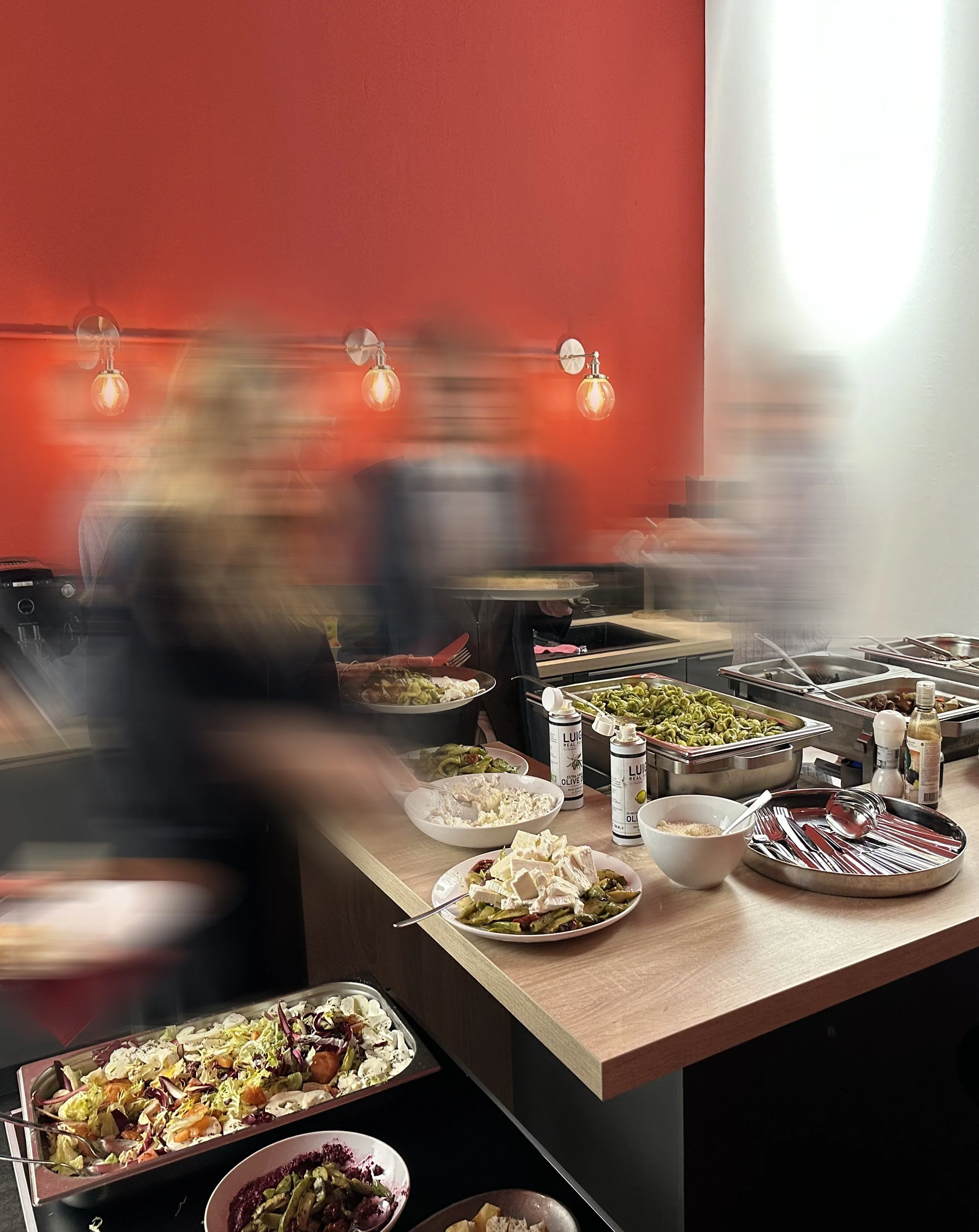Buffet mit Salaten und warmen Speisen in einer modernen Küche, einige Gäste bedienen sich, im Hintergrund eine rote Wand mit Wandlampen und mehreren Menschen in Bewegung.