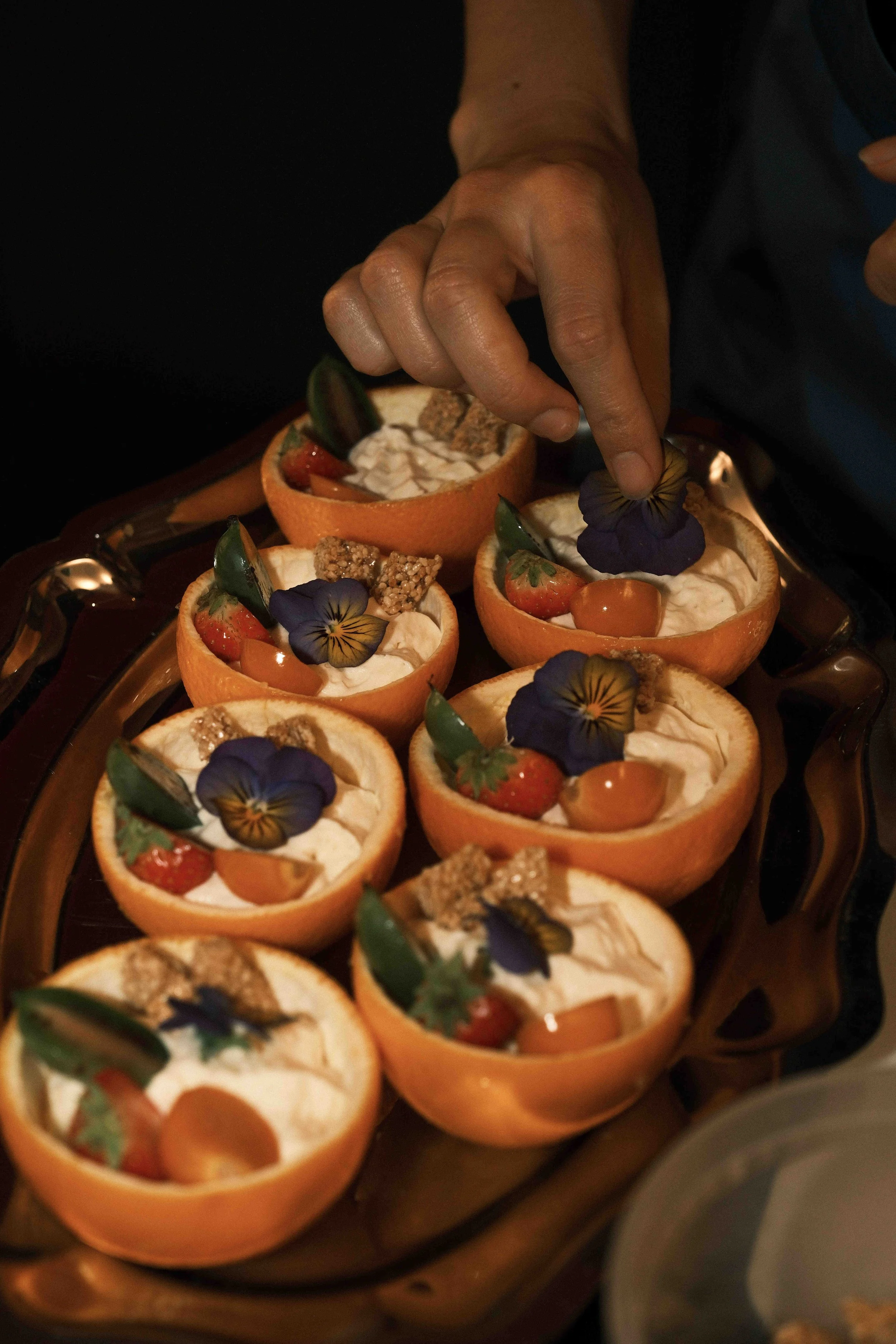 Hände, die kleine Orangenhälften mit cremiger Füllung, Beeren, essbaren Blumen und Dekorationen garnieren.