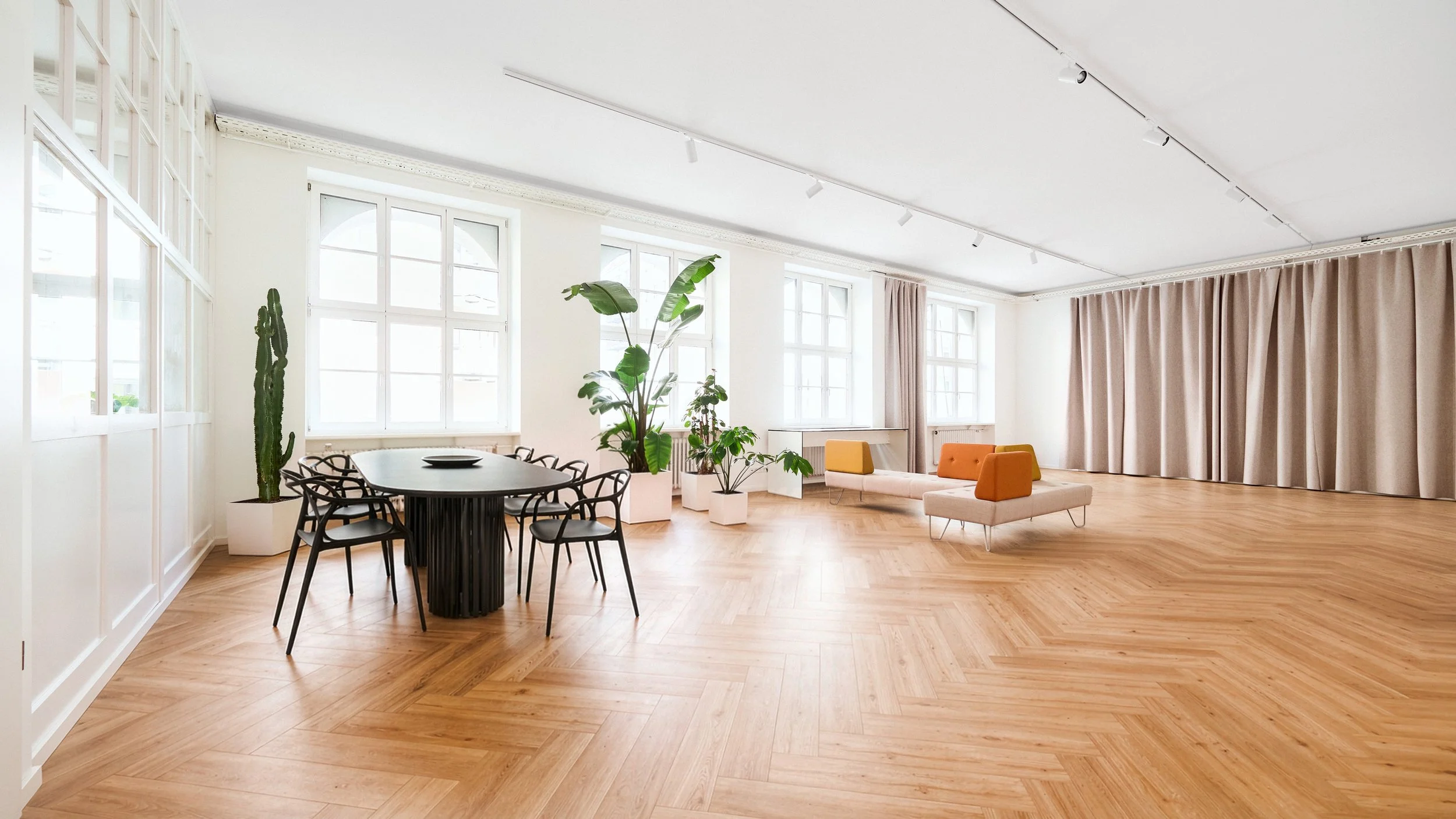 Ein heller, moderner Raum mit großen Fenstern, Holzboden, mehreren Pflanzen, einem runden Esstisch mit schwarzen Stühlen und einem Sitzbereich mit orangefarbenen und beige Sofas, Vorhängen an der Wand.