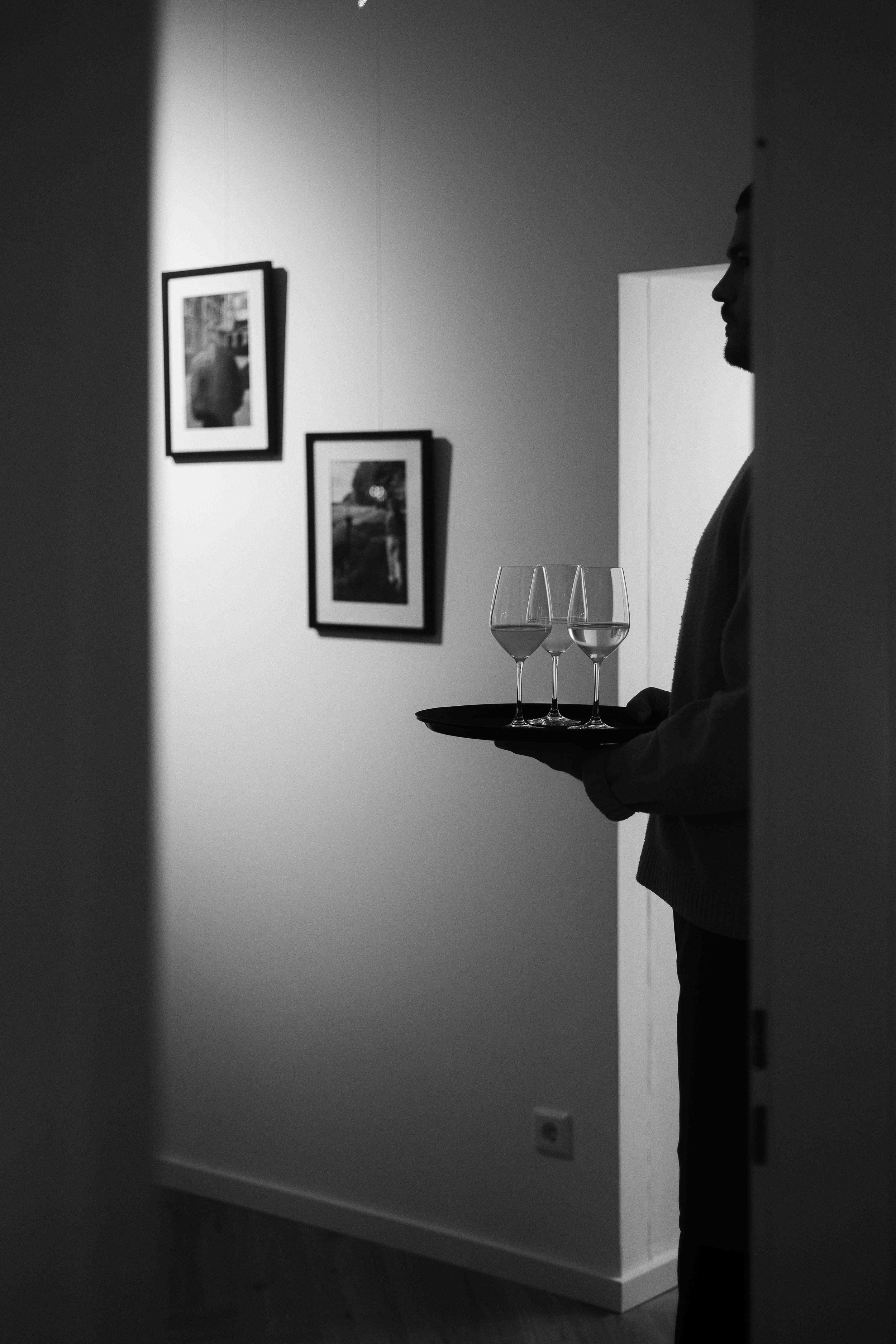 Ein Mann hält einen Tablett mit drei Weingläsern, die gefüllt sind, in einem Raum mit hellen Wänden und an der Wand hängen Fotos. Das Bild ist in Schwarz-Weiß gehalten.