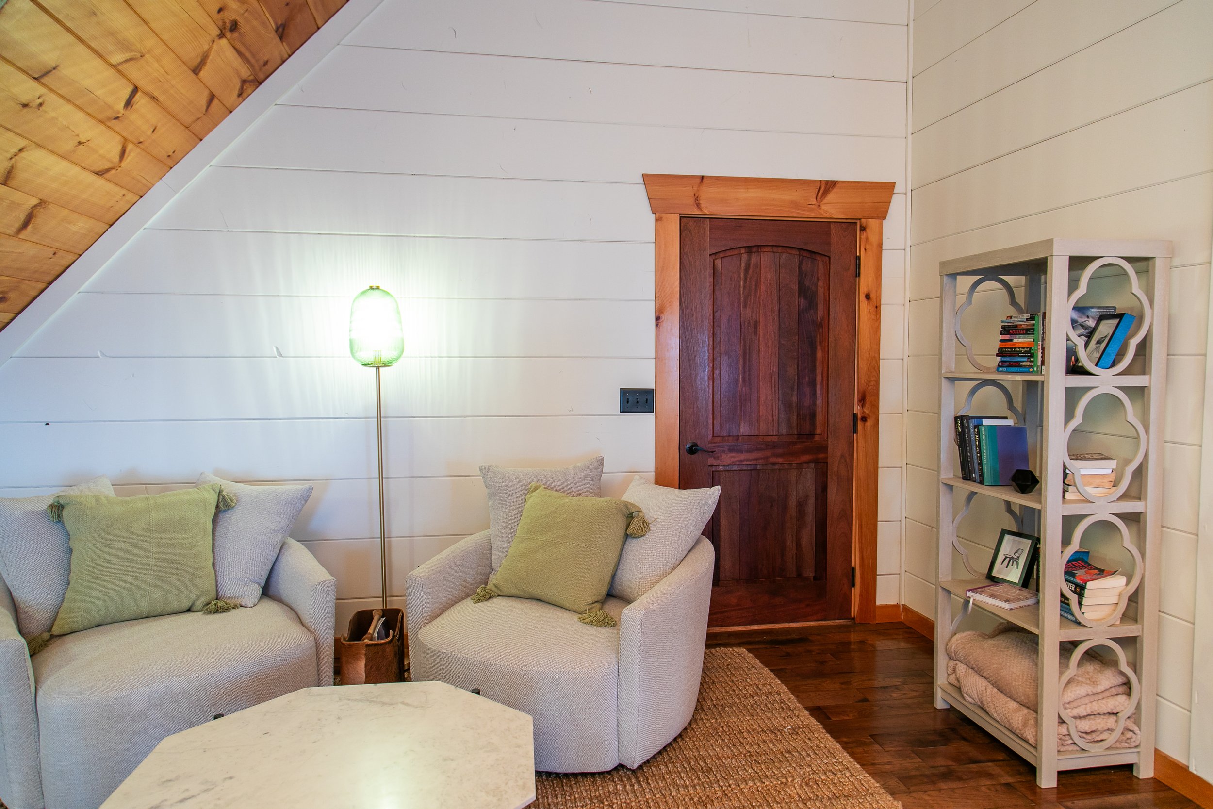 Living room with two beige armchairs with green pillows, a wooden door, a white bookcase with books and blankets, a textured rug, and a floor lamp with a green shade.