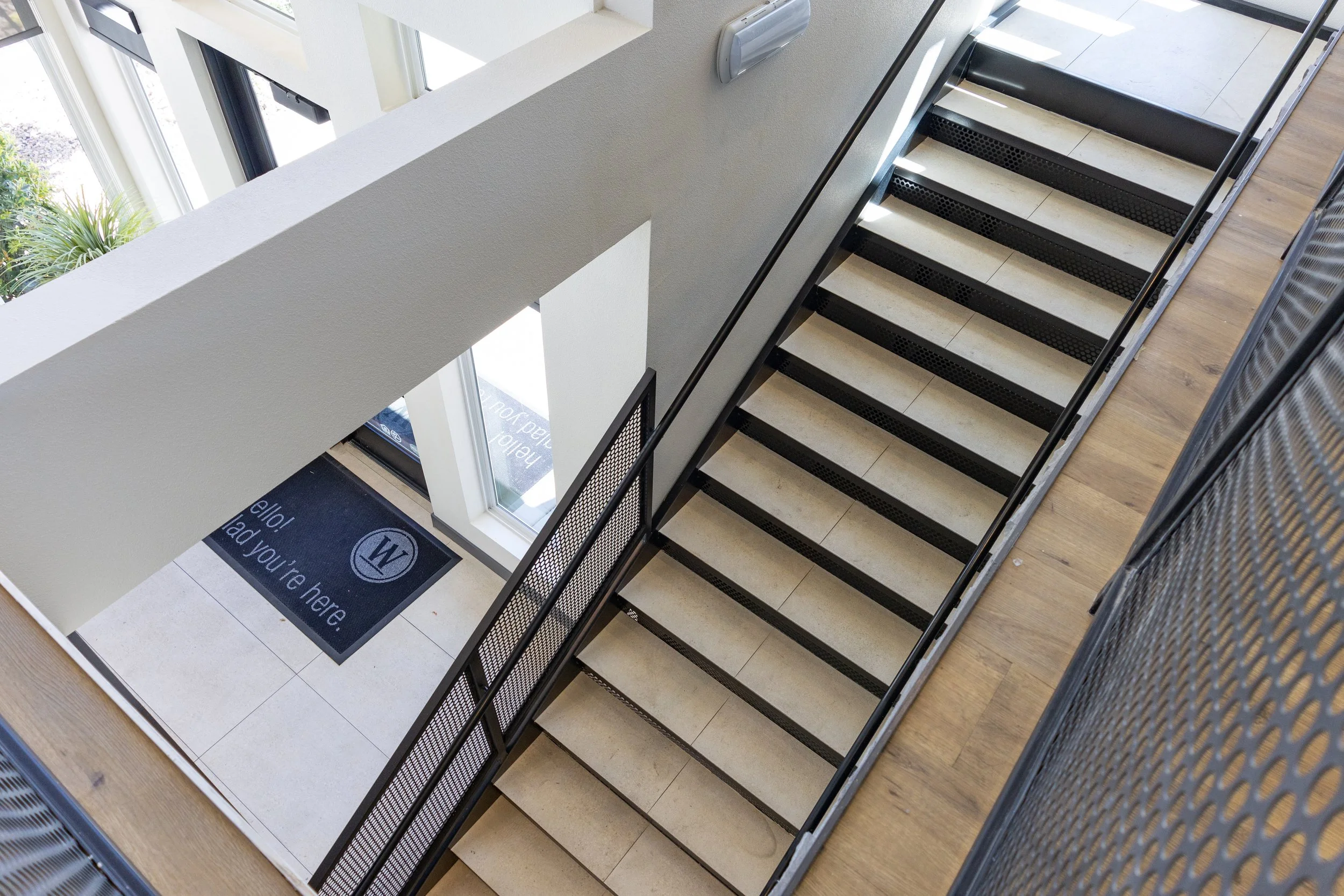 View of a staircase from above in a modern building, with a black metal railing and light-colored steps, next to a wall with large windows and a welcome mat at the entrance.