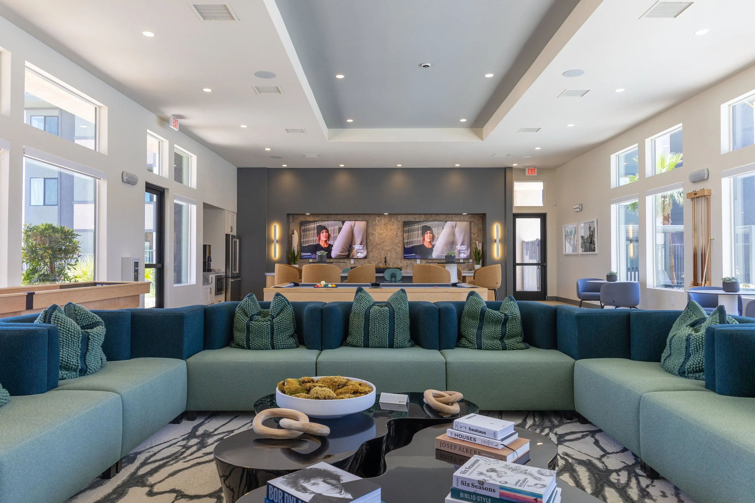 Modern, spacious lounge area with a large curved green and blue sofa, a black coffee table with books and decorative objects, and a wall with two flat-screen televisions. The room has large windows, natural light, and contemporary decor.