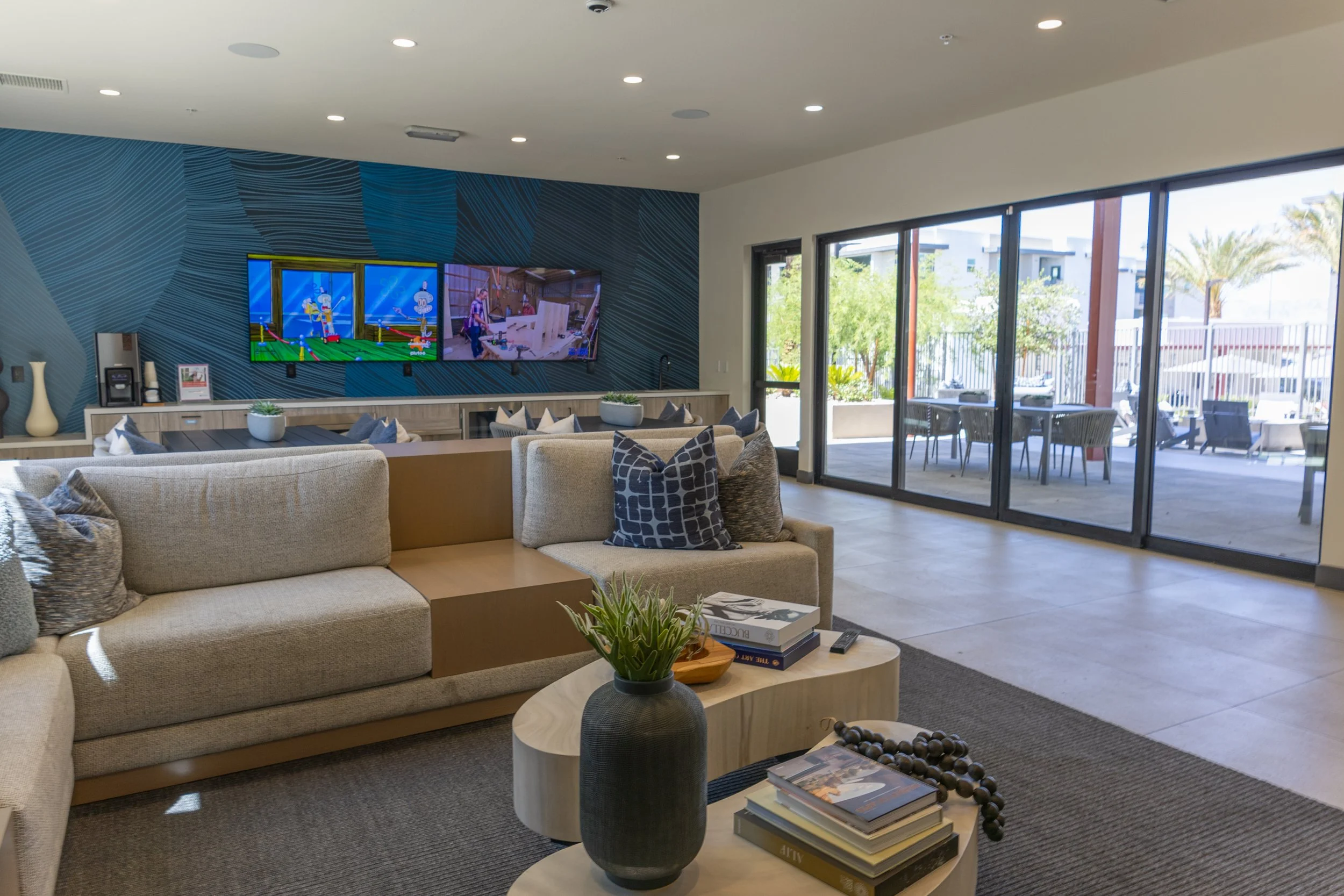 Modern living room with beige sectional sofa, throw pillows, wooden coffee table with books, vase with plant, view of outdoor patio with dining table, and two TV screens on textured blue wall.