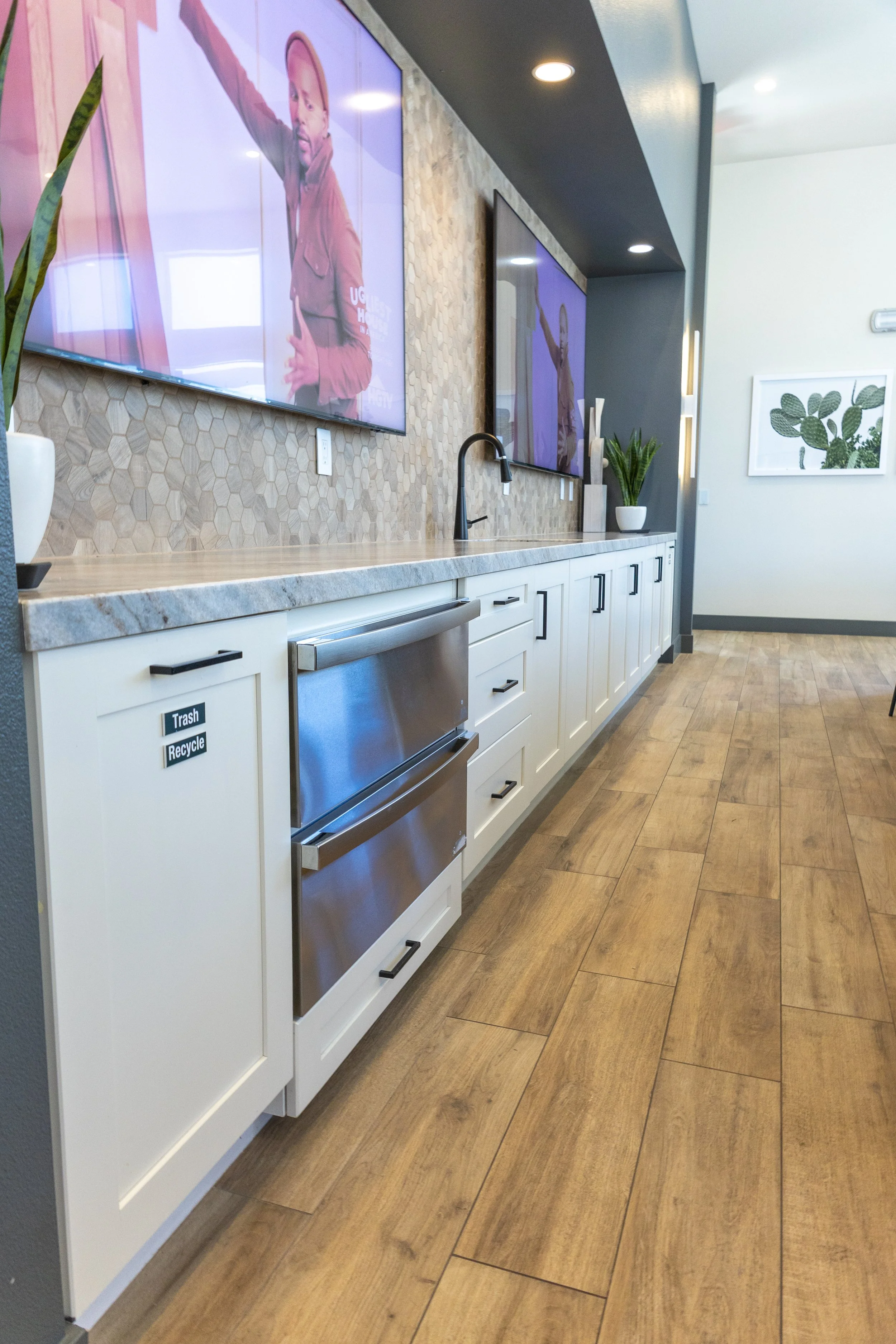 Modern kitchenette with white cabinets, wood-look flooring, wall-mounted TVs, decorative plants, and a honeycomb tile backsplash.
