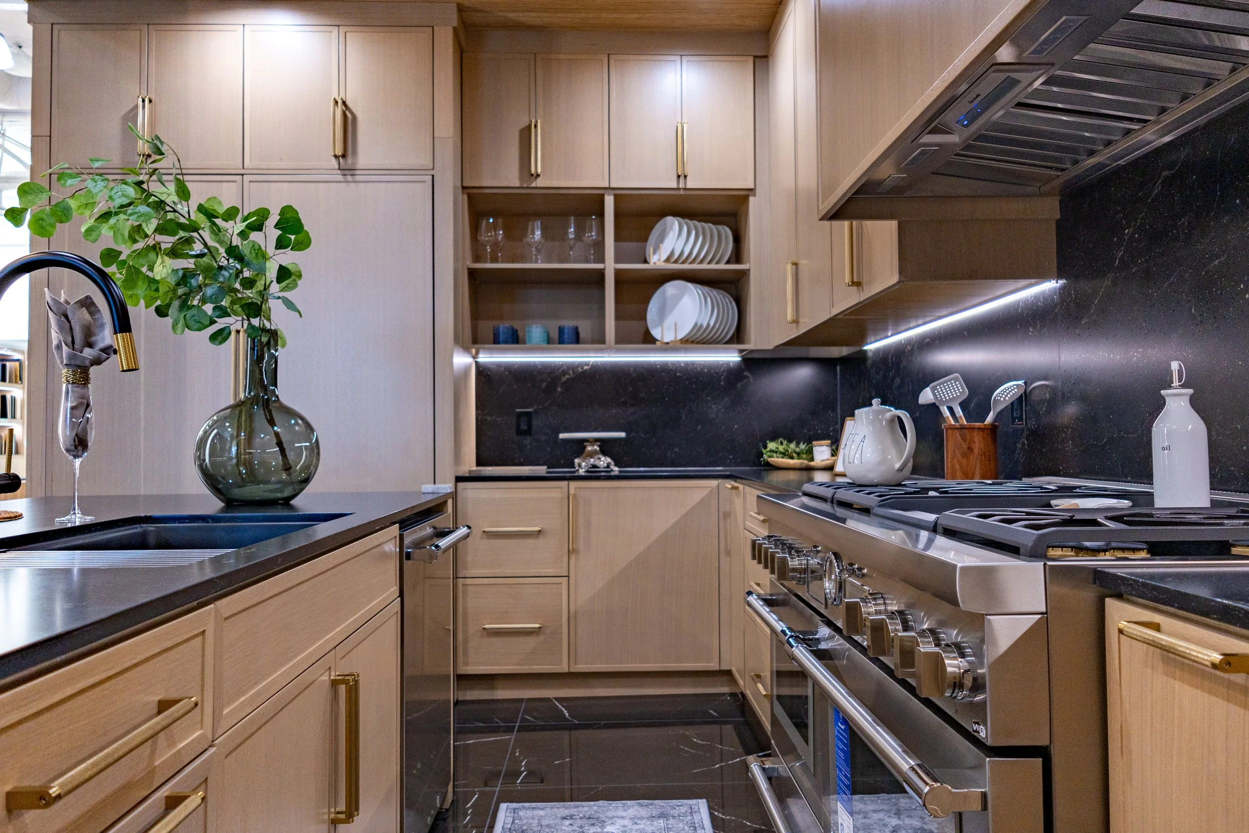 A modern kitchen with light wood cabinets, black marble countertops, and stainless steel appliances. There is a vase with green foliage on the counter, open cabinet with dishes and glasses, and various kitchen utensils.
