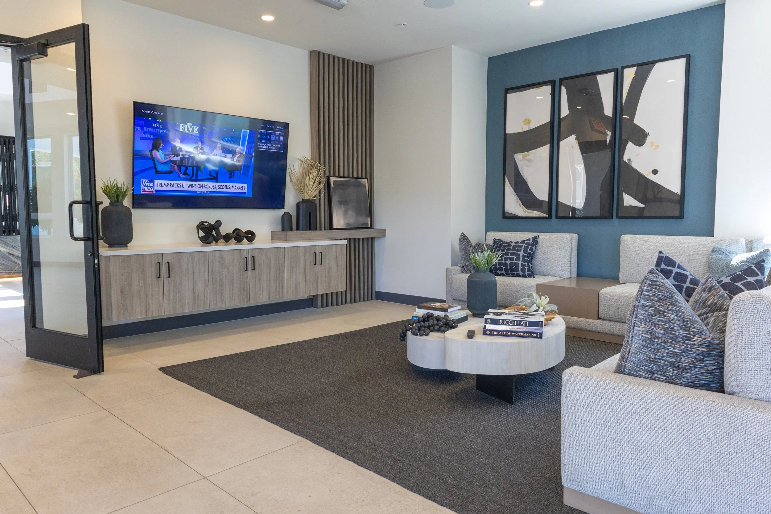 Modern living room with a wall-mounted TV, abstract artwork, a beige sofa with patterned pillows, a black coffee table with books and decorative items, a dark area rug, and a sideboard with vases and sculptures.