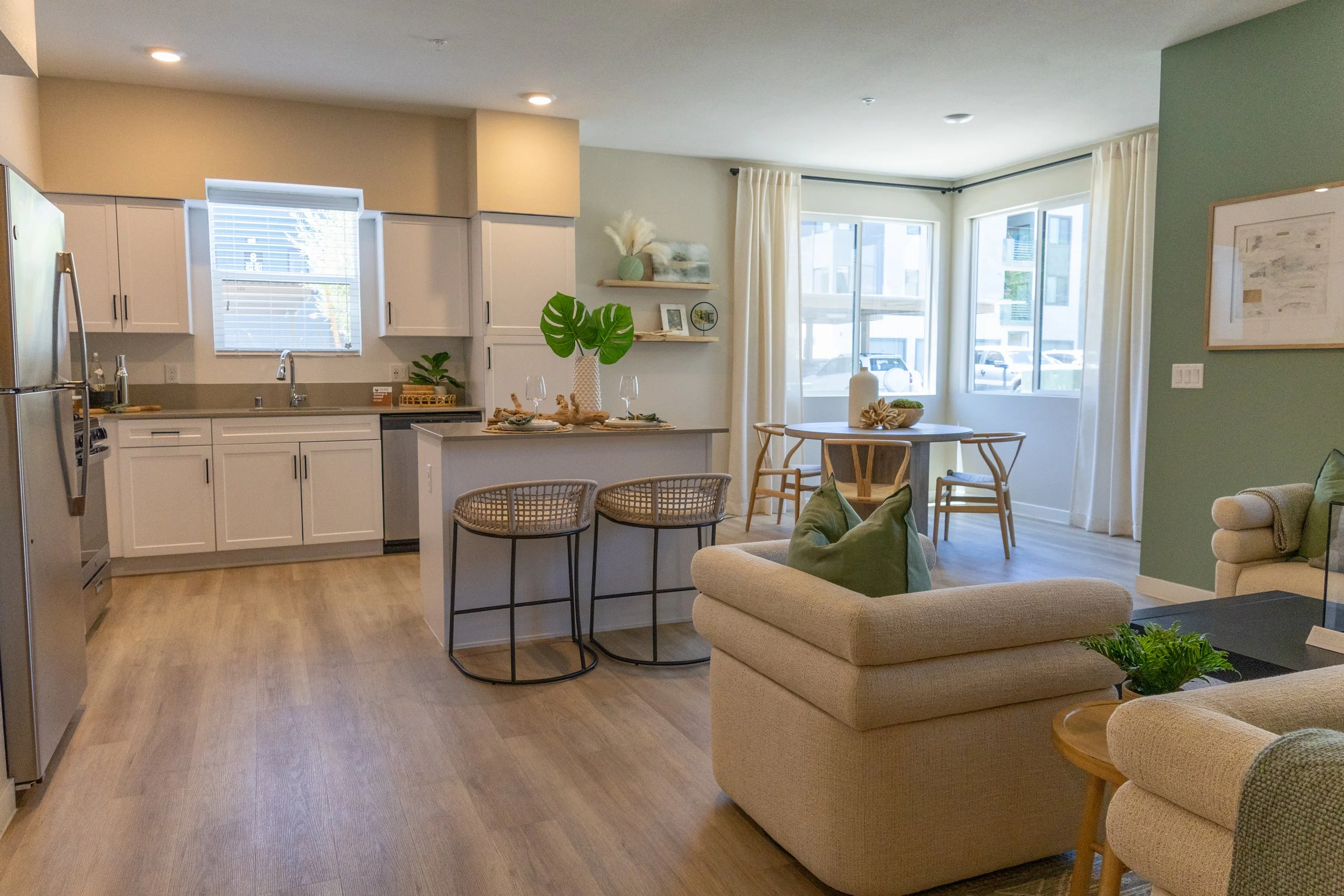 Open-concept living room and kitchen with white cabinets, beige walls, green accent wall, wooden floors, and large windows with white curtains, decorated with plants and minimalist decor.