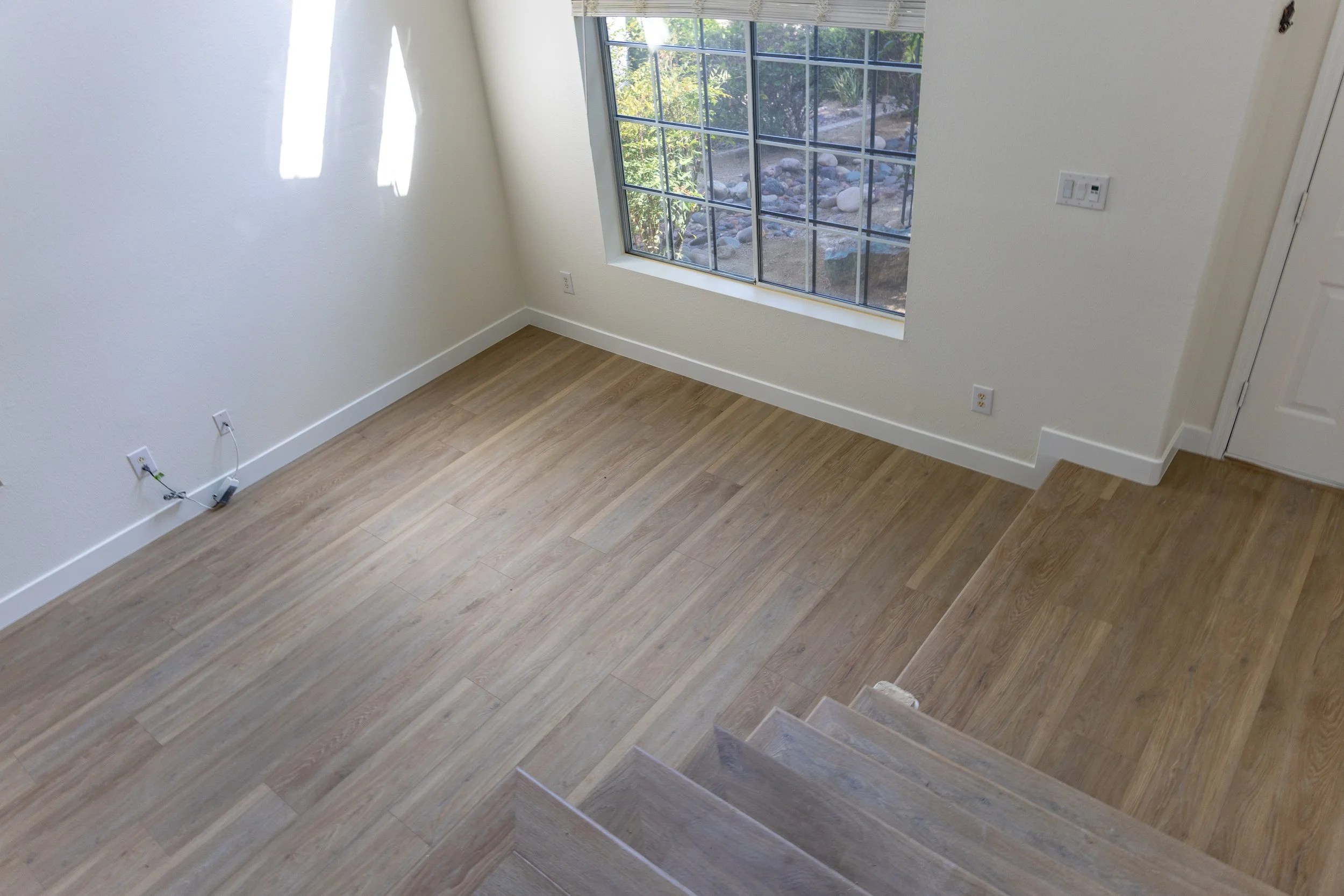 Empty room with hardwood flooring, white walls, a large window with grid panes, and a staircase leading down to the room.