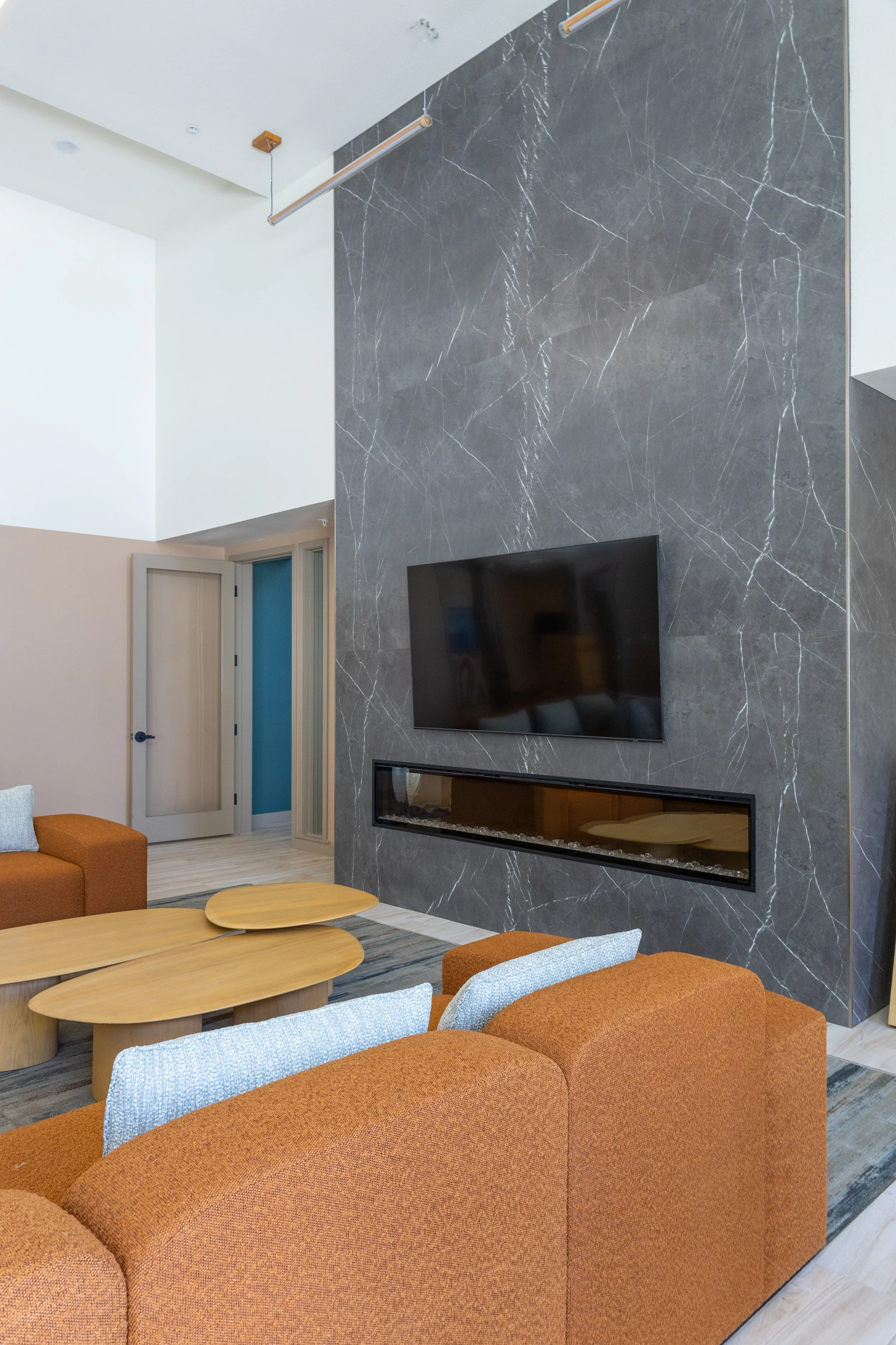 Living room with a black marble wall, mounted flat-screen TV, glass-enclosed fireplace, orange couches with gray pillows, and wooden coffee tables.