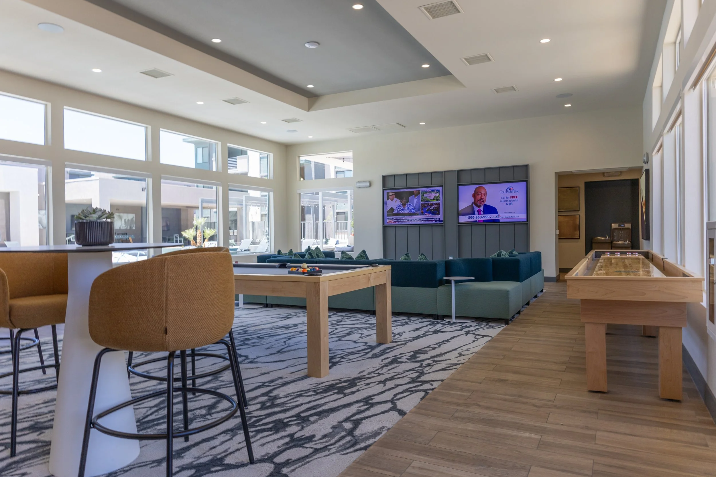 A spacious lounge area with large windows, a TV wall, and seating including bar stools and a green sectional sofa, with a carpet and a wooden side table.