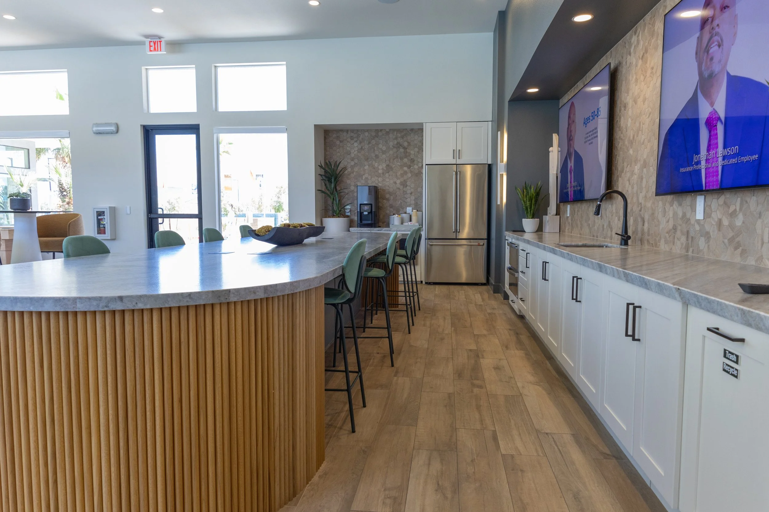 Modern kitchen and lounge area with a curved island, bar stools, stainless steel refrigerator, and two wall-mounted TVs.