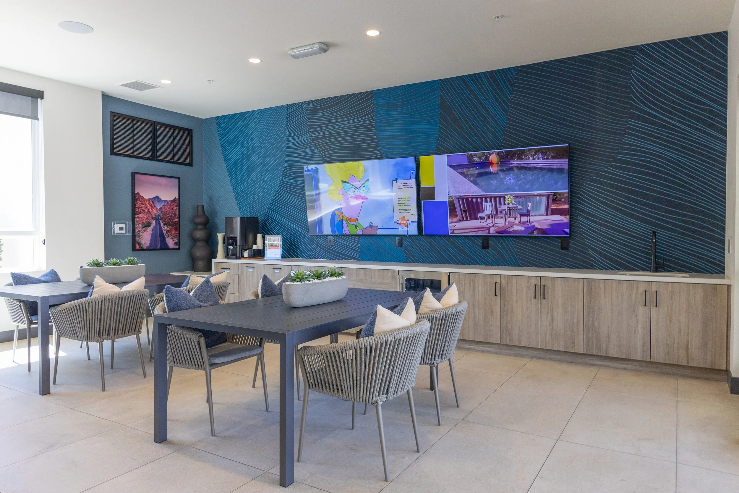 Modern lounge area with two large flat-screen TVs on a patterned blue accent wall, a dark wooden table surrounded by gray chairs with cushions, and decor including potted plants and artwork, near a window with natural light.