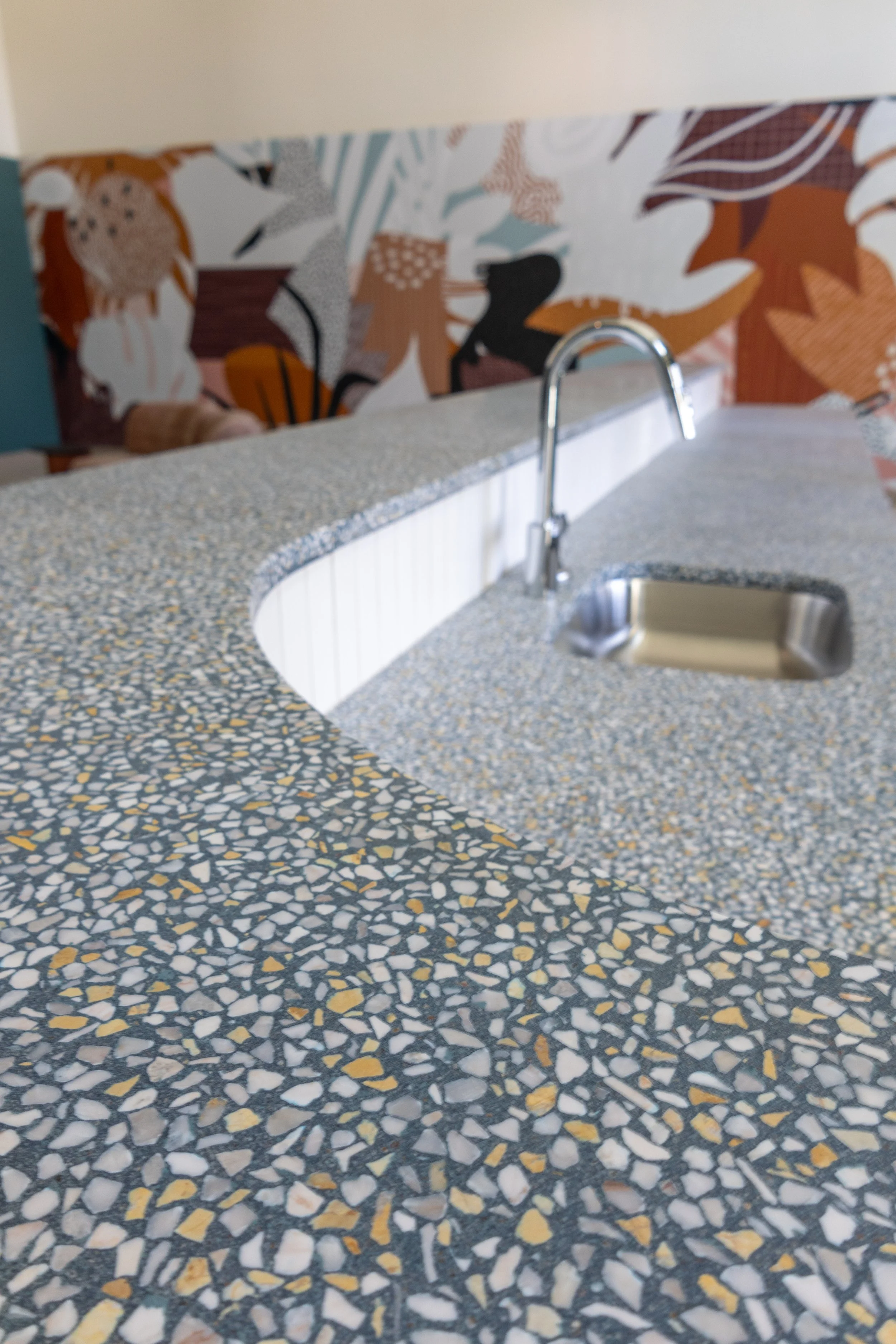 Close-up of a mosaic countertop with a built-in sink and chrome faucet in a kitchen or bar area, with a colorful abstract mural in the background.