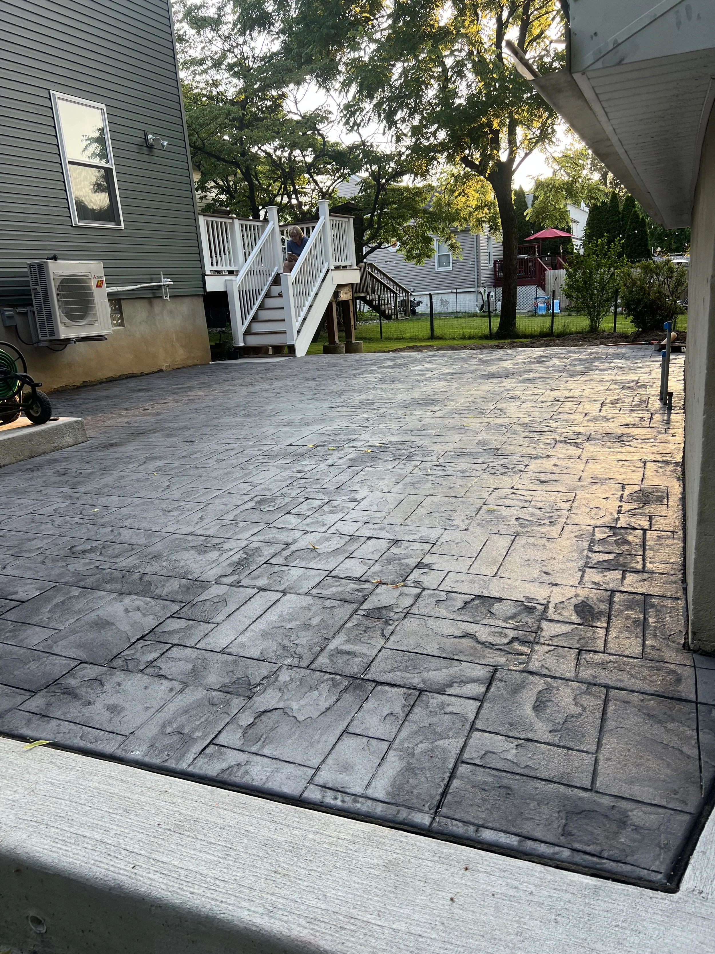 View of a backyard with stamped concrete patio, neighboring houses, trees, and a person on the deck stairs