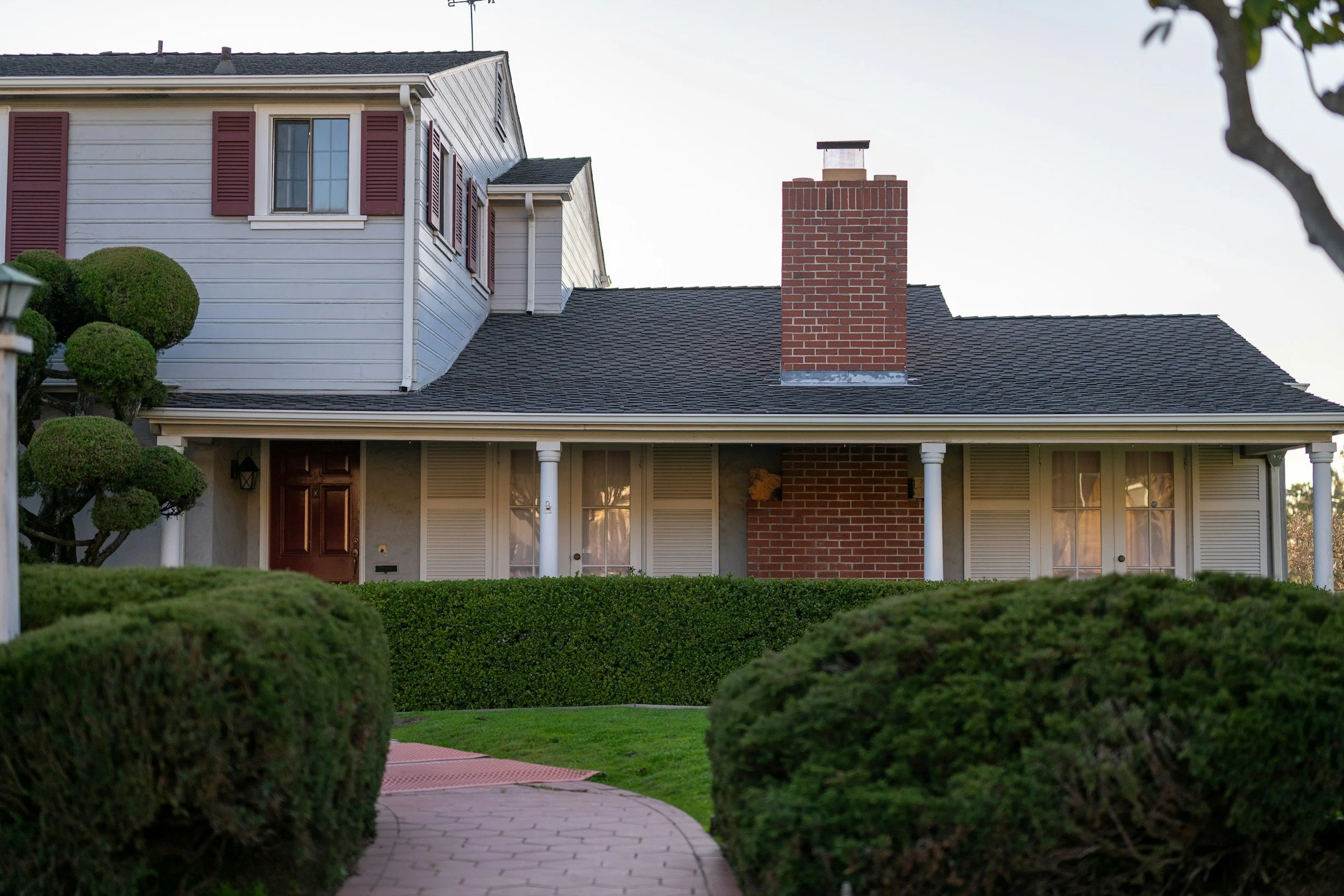 Front view of a two-story house with white siding, red shutters, brick chimney, and a front porch with columns, surrounded by trimmed bushes and a winding brick walkway.