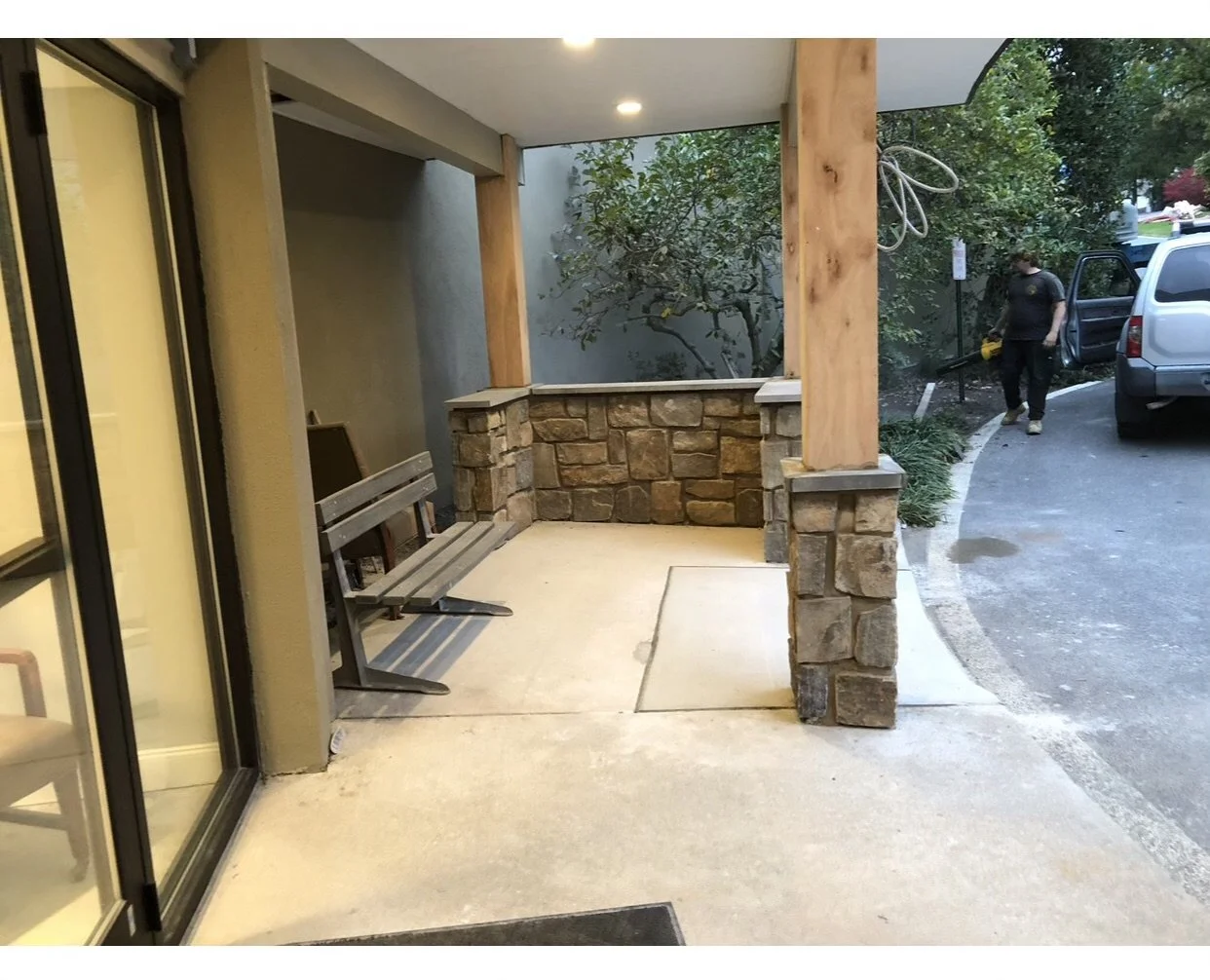 Front porch of a building with a brick and wood structure, a bench, and a sidewalk. A woman is walking near a parked SUV with an open door. Trees and greenery are in the background.