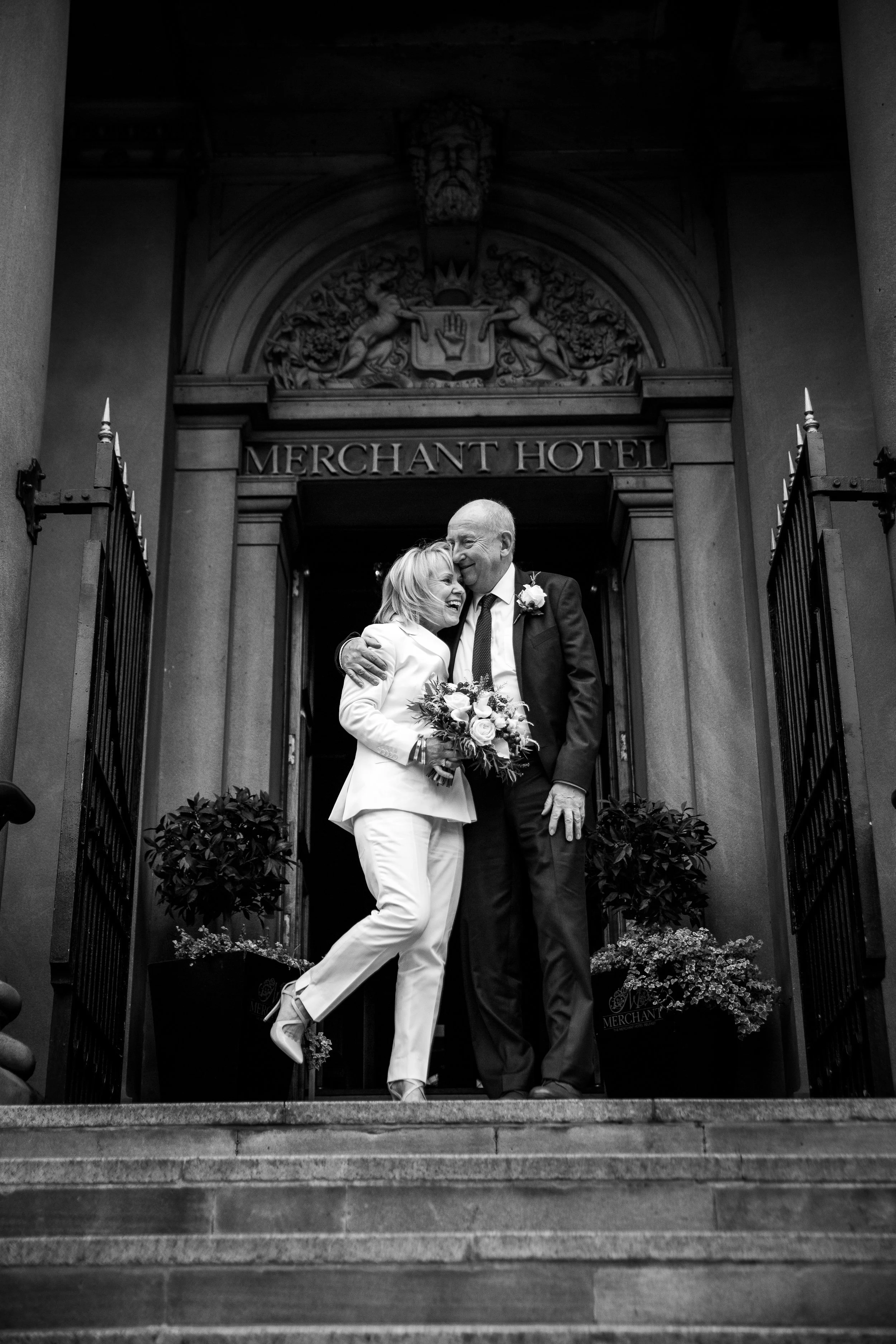 A smiling woman and an older man embracing each other and sharing a laugh on the steps of the Merchant Hotel, with an ornate sign and lion sculptures in the background.