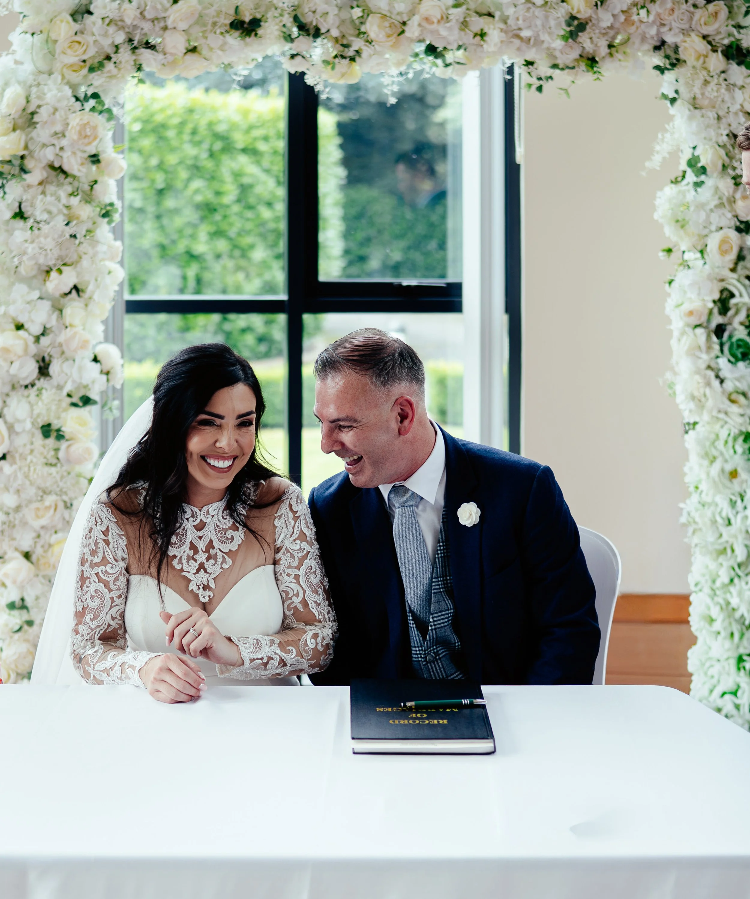 A bride and groom holding hands in a park with yellow flowers and bare trees in the background.
