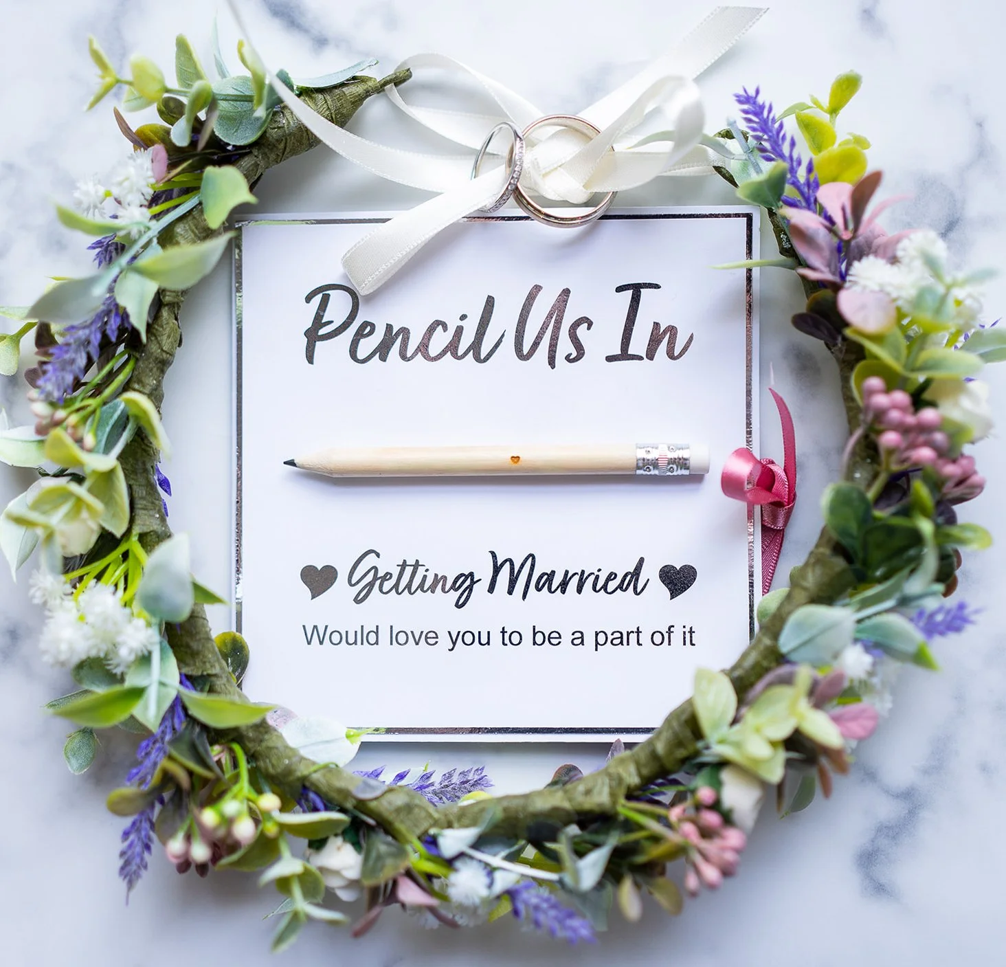 A floral wreath surrounds a card with the words 'Pencil Us In' and 'Getting Married' written on it, with a pencil placed underneath and two wedding rings attached to a satin ribbon at the top.