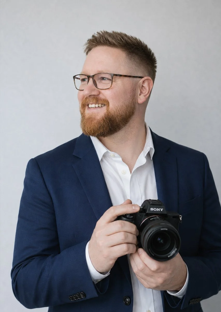 A man with glasses, a beard, and short hair wearing a dark blue suit and white shirt, holding a Sony camera, smiling and looking to the side.