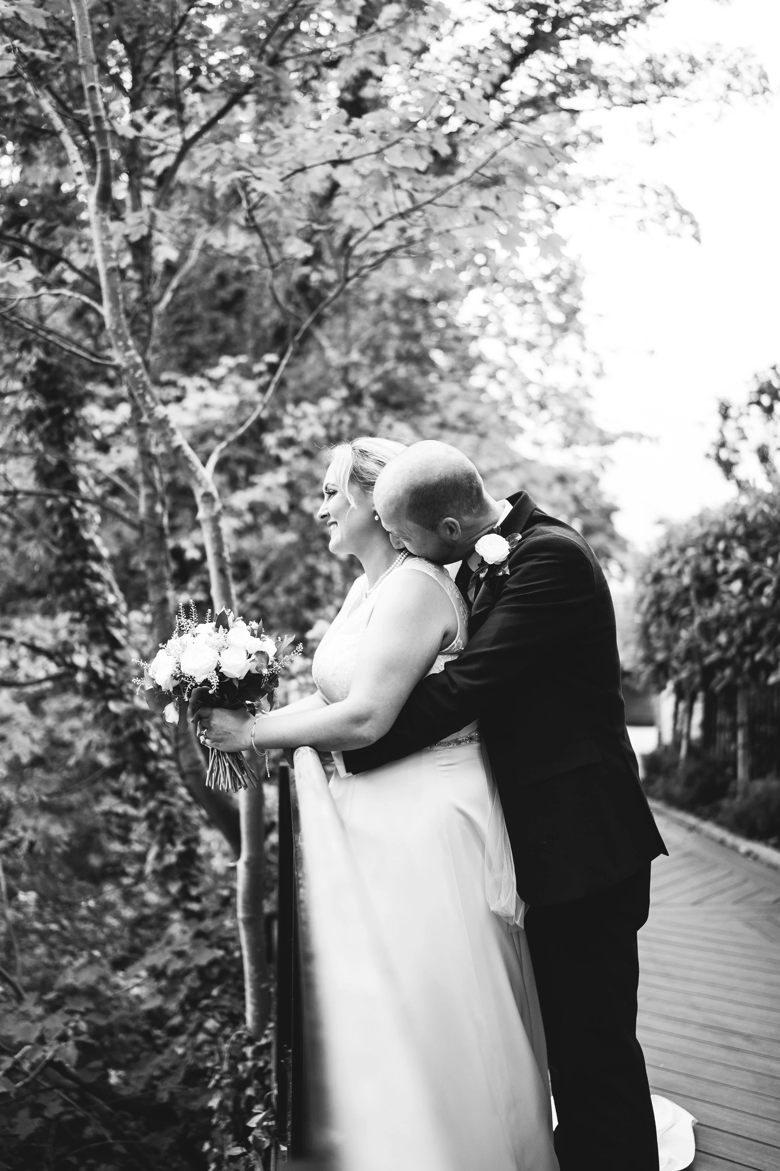 A black and white photo of a bride and groom outdoors. The groom, dressed in a suit, is kissing the bride on the cheek from behind. The bride is smiling, holding a bouquet of flowers, and wearing a wedding dress. They are standing next to a wooden railing with trees and foliage in the background.