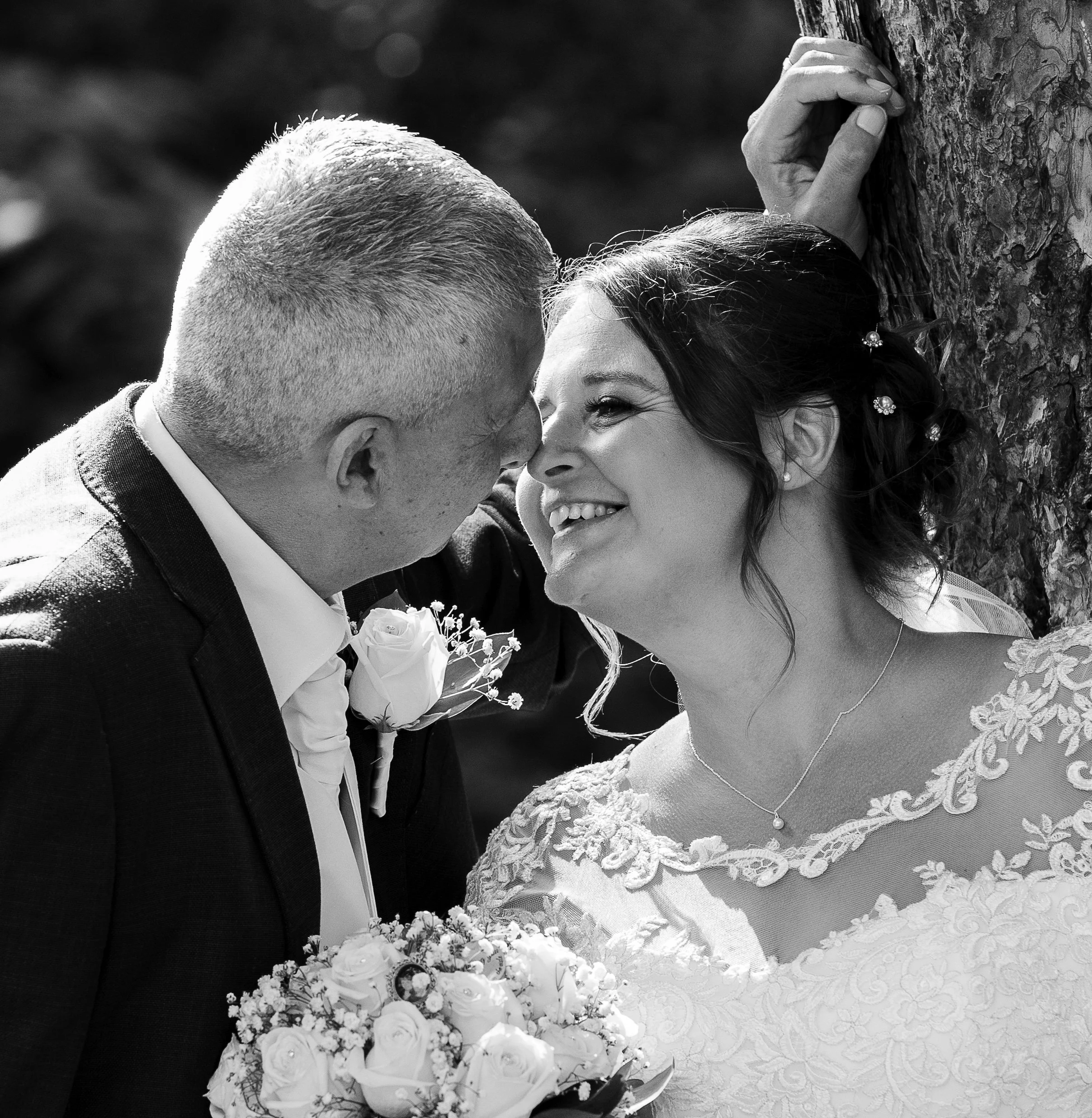 A black-and-white photo of a smiling bride and groom touching noses, holding a bouquet of white flowers, with a textured tree trunk in the background.