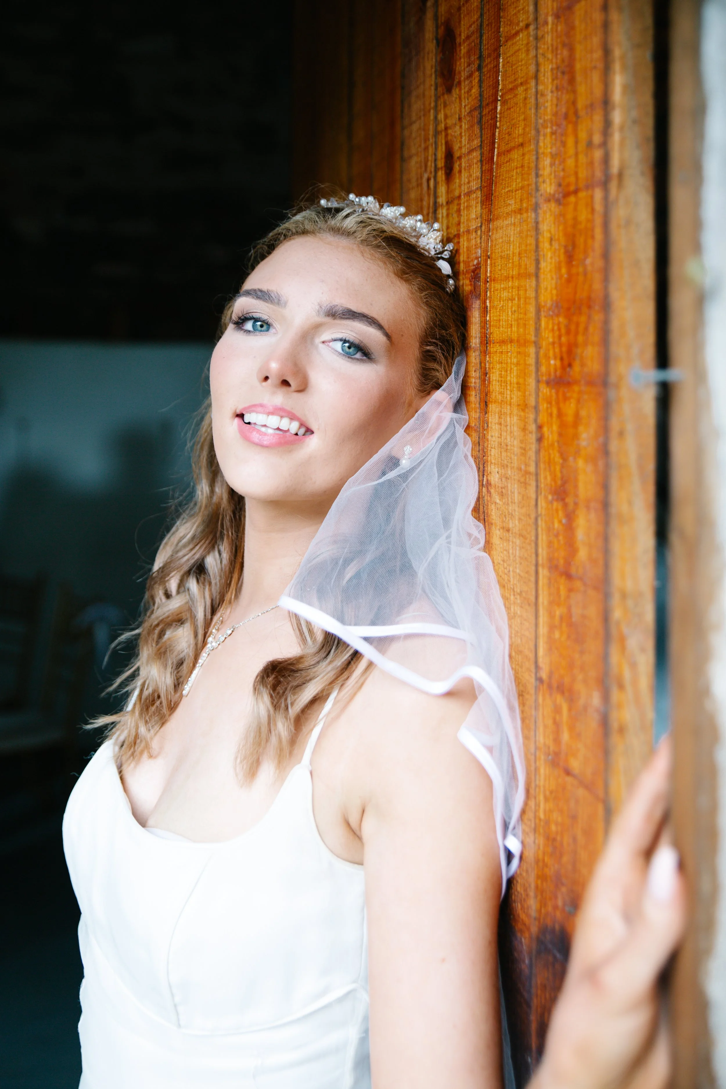 A young woman dressed in a white wedding gown with a lace veil and pearl accessories, standing next to a wooden wall.