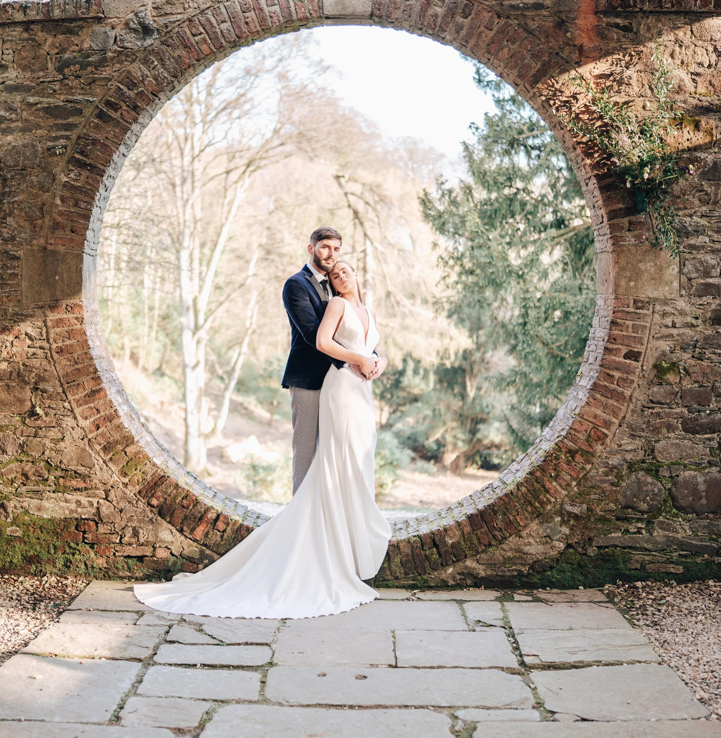 A couple in wedding attire standing under an arched brick structure with trees in the background, sunlight filtering through the trees.