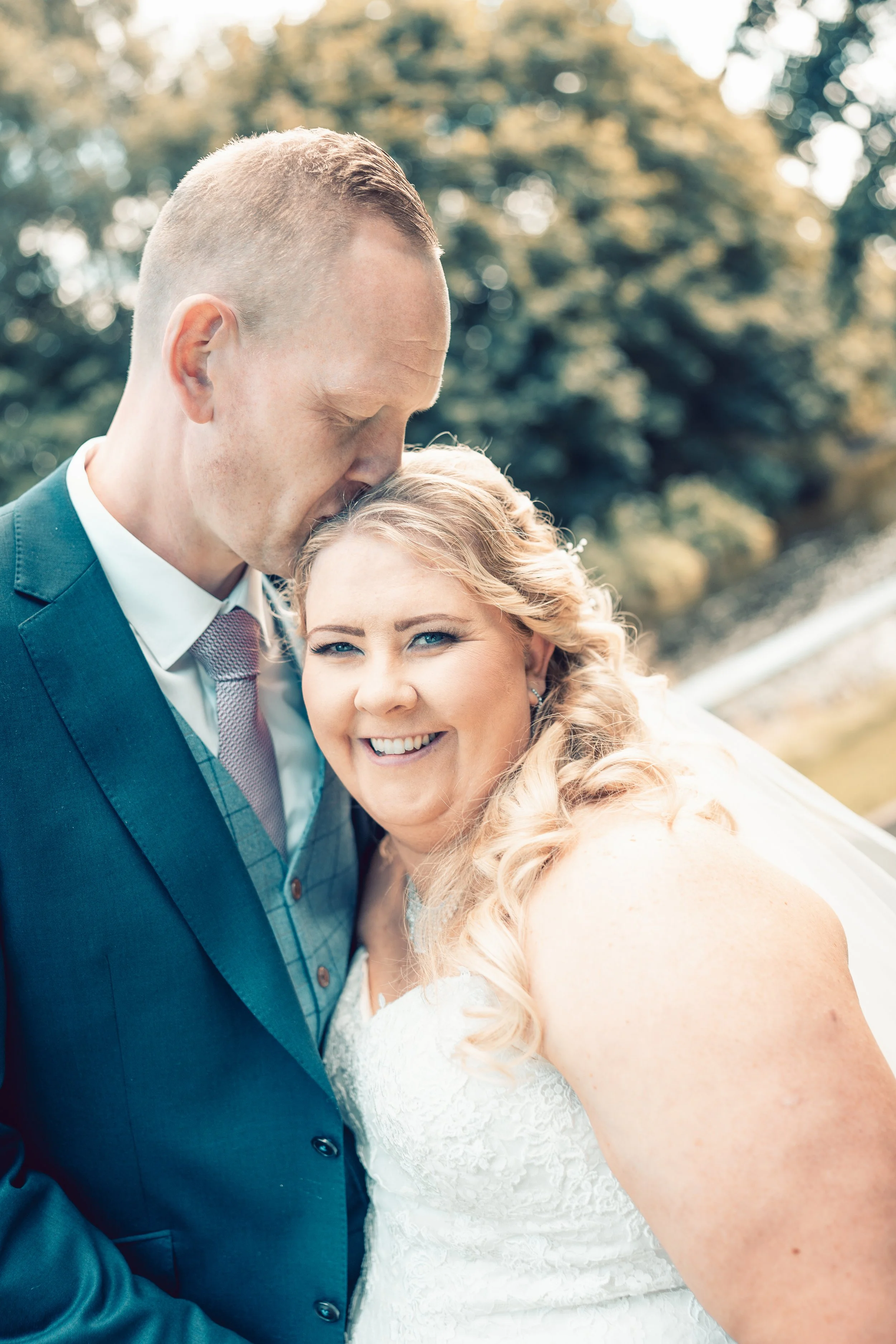 A bride and groom on their wedding day, outdoors with blurred trees in the background. The groom is kissing the bride's forehead, and she is smiling happily.