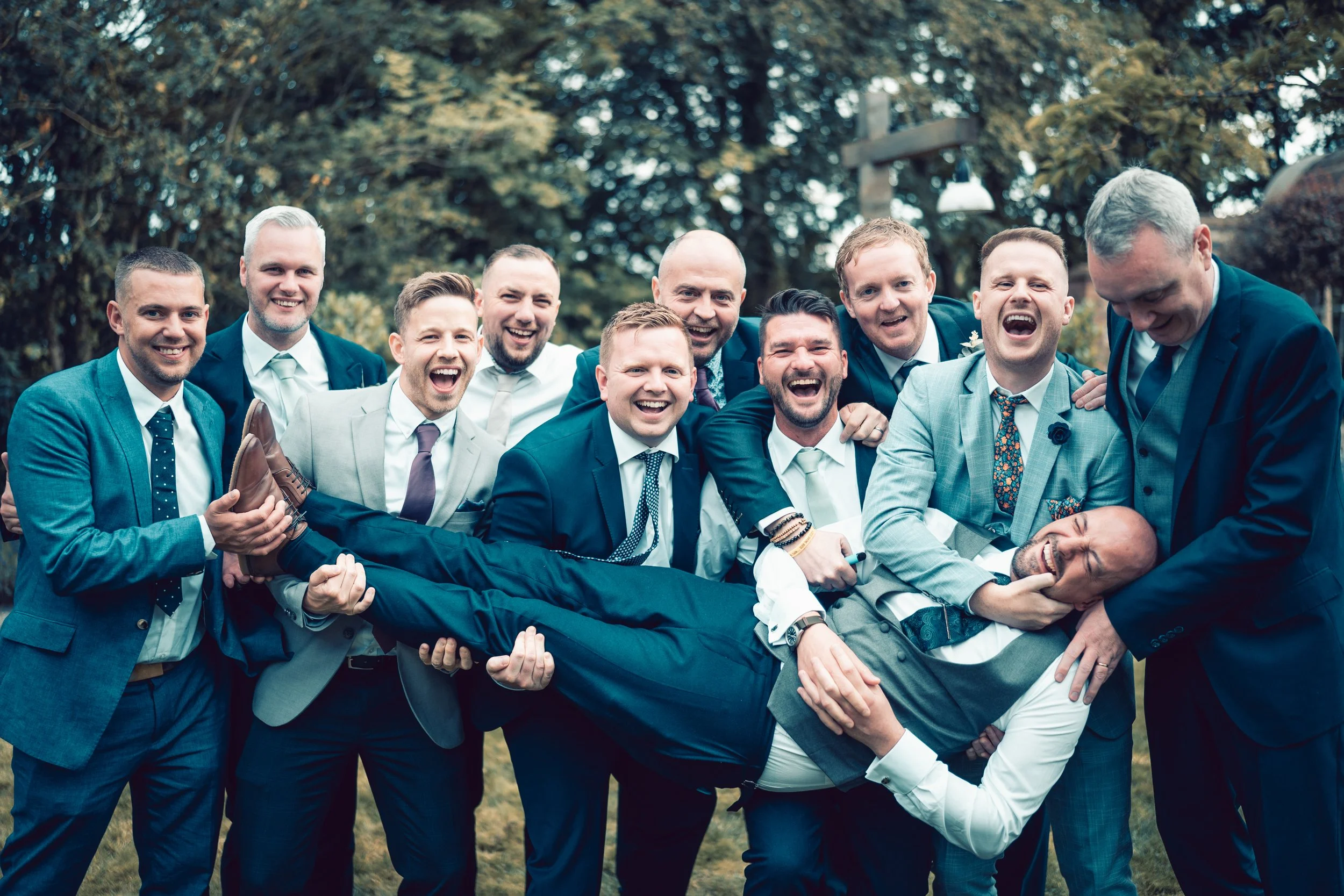 A group of eleven men dressed in formal suits laughing and playing together outdoors.