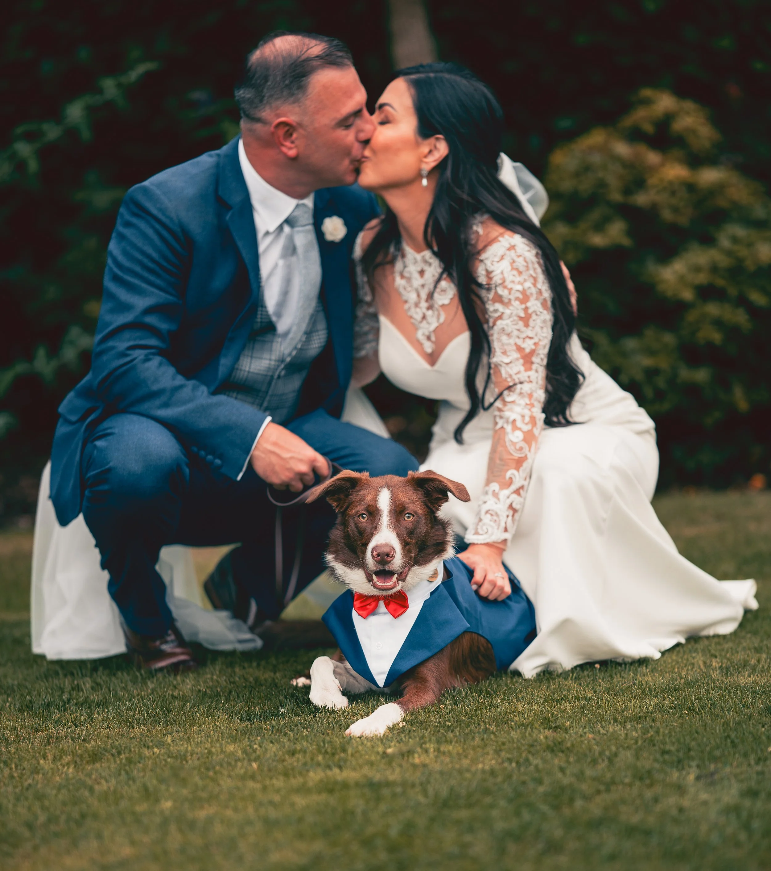 A couple sharing a kiss on their wedding day, with the bride sitting on the grass and the groom leaning in, a dog with a red bowtie lying in front of them.
