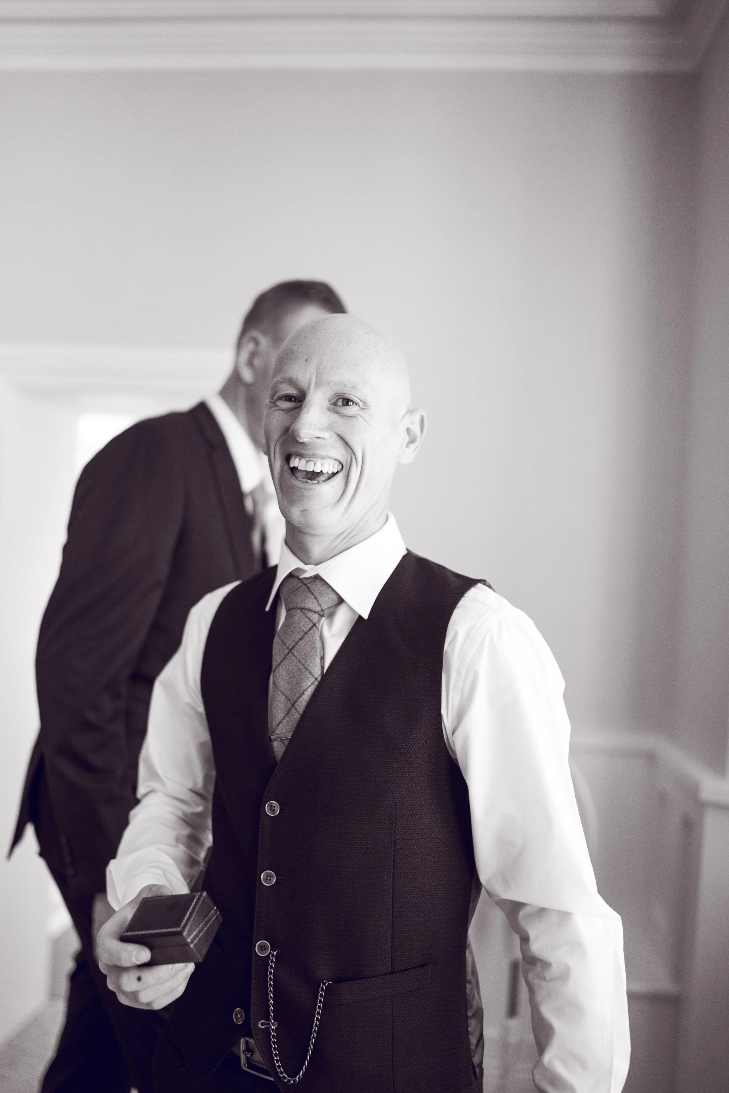 A man with a bald head and a suit smiling and holding a small box, standing in front of another man, both in formal attire, in an indoor setting.