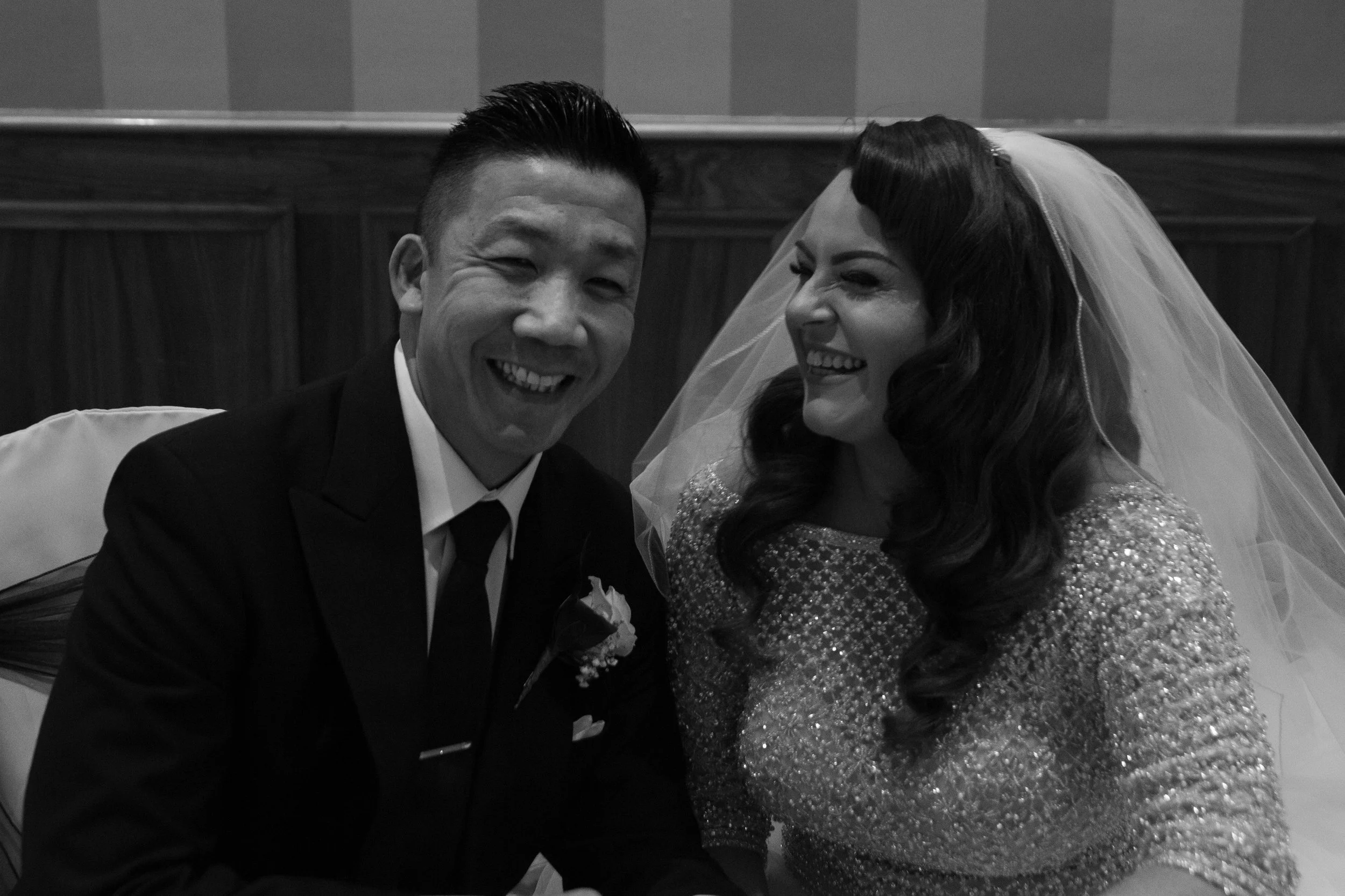 A happy couple, the man in a tuxedo and the woman in a bridal gown with a veil, smiling and celebrating at their wedding.