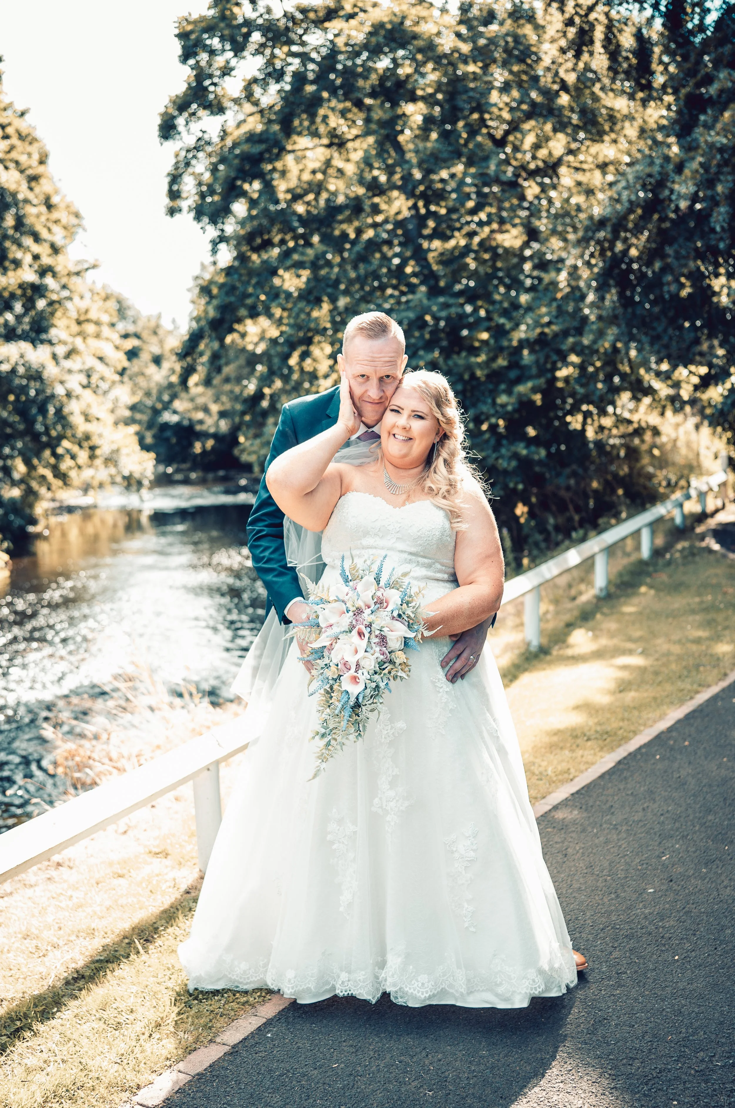 A bride holding a bouquet of flowers with a groom standing behind her, outdoors near a waterway and surrounded by trees.
