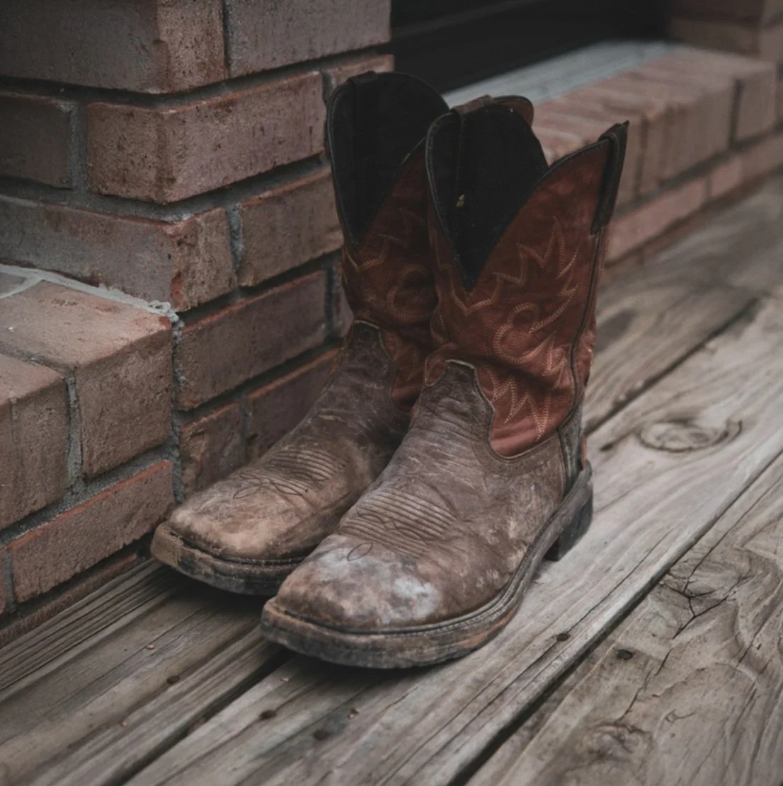 A pair of dirty cowboy boots leaning against a brick wall on a wooden floor.