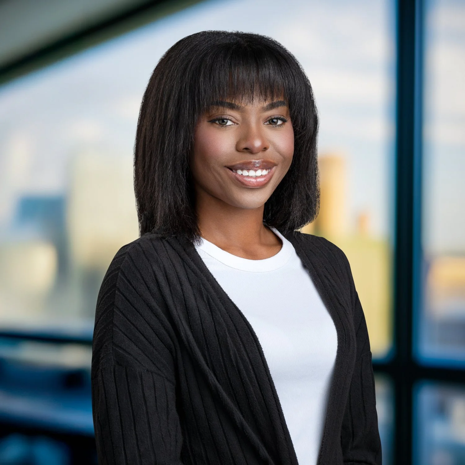 A smiling African American woman with shoulder-length black hair, wearing a white shirt and black cardigan, standing indoors with blurred cityscape in the background.