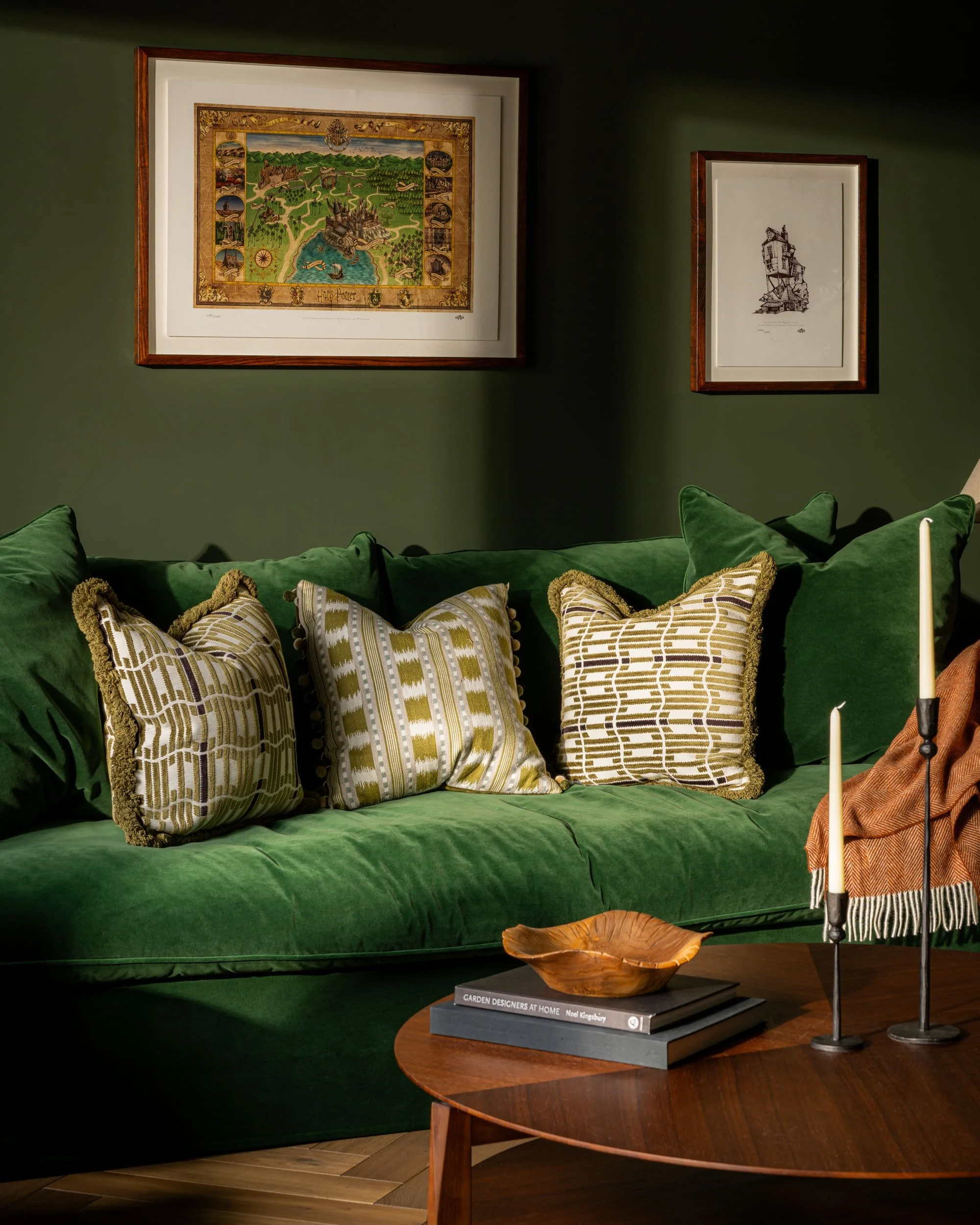 Living room with a green velvet sofa decorated with multiple patterned pillows, green walls with two framed pictures, a wooden coffee table with a decorative bowl and a book titled 'Garden Designers at Home', and a candle holder with candles.